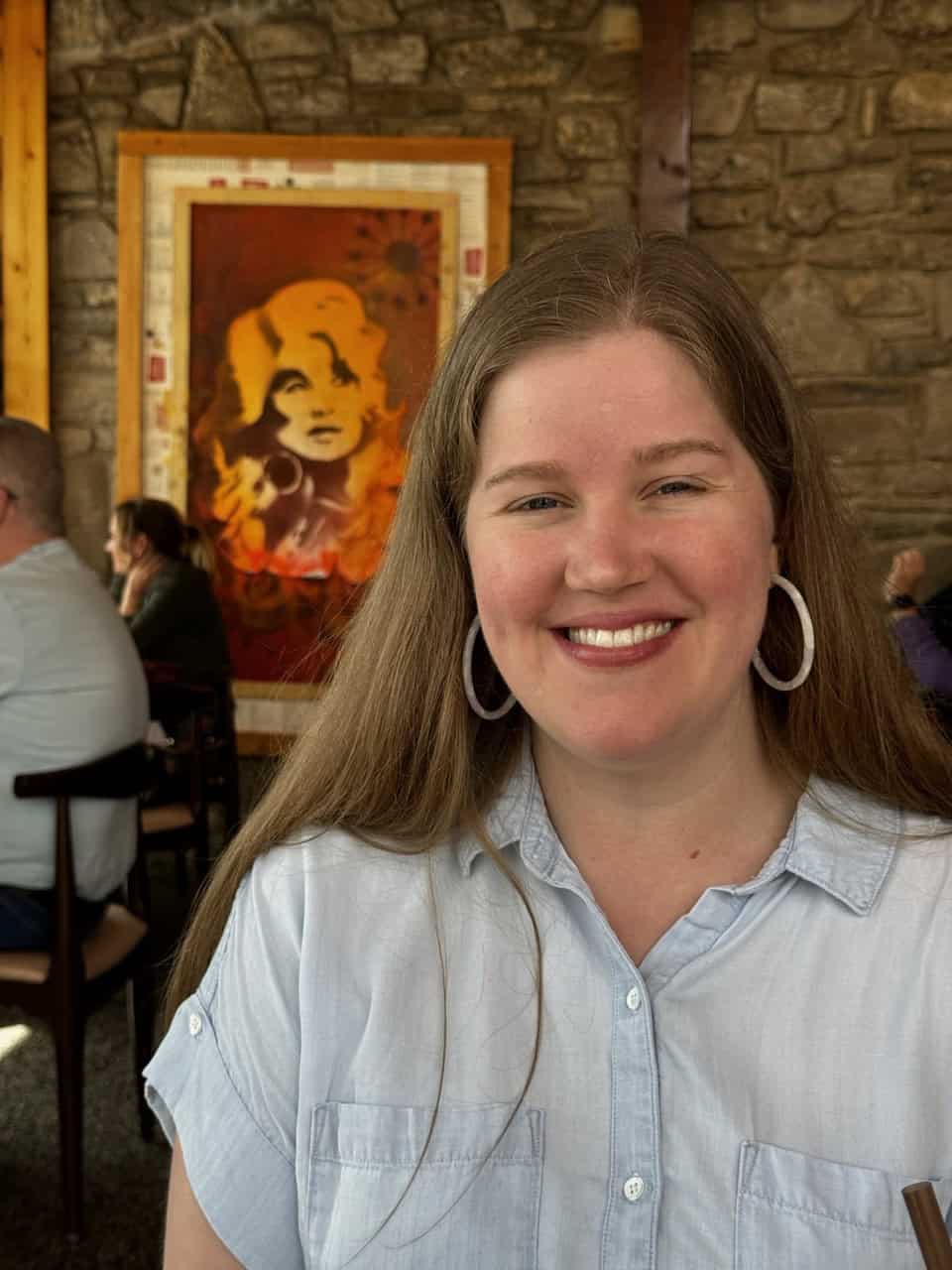 A smiling woman with long hair and hoop earrings sits indoors in front of a stone wall adorned with Nashville-inspired art.