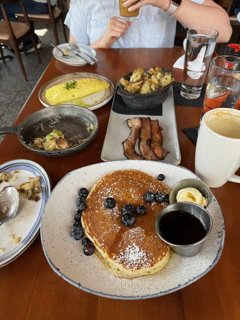 A table set for a Nashville breakfast features pancakes topped with blueberries, an omelette, crispy bacon strips, and a savory potato dish. Nearby rest a mug of steaming coffee and a glass of water. A person is seated, partially visible, savoring the moment with their cup in hand.