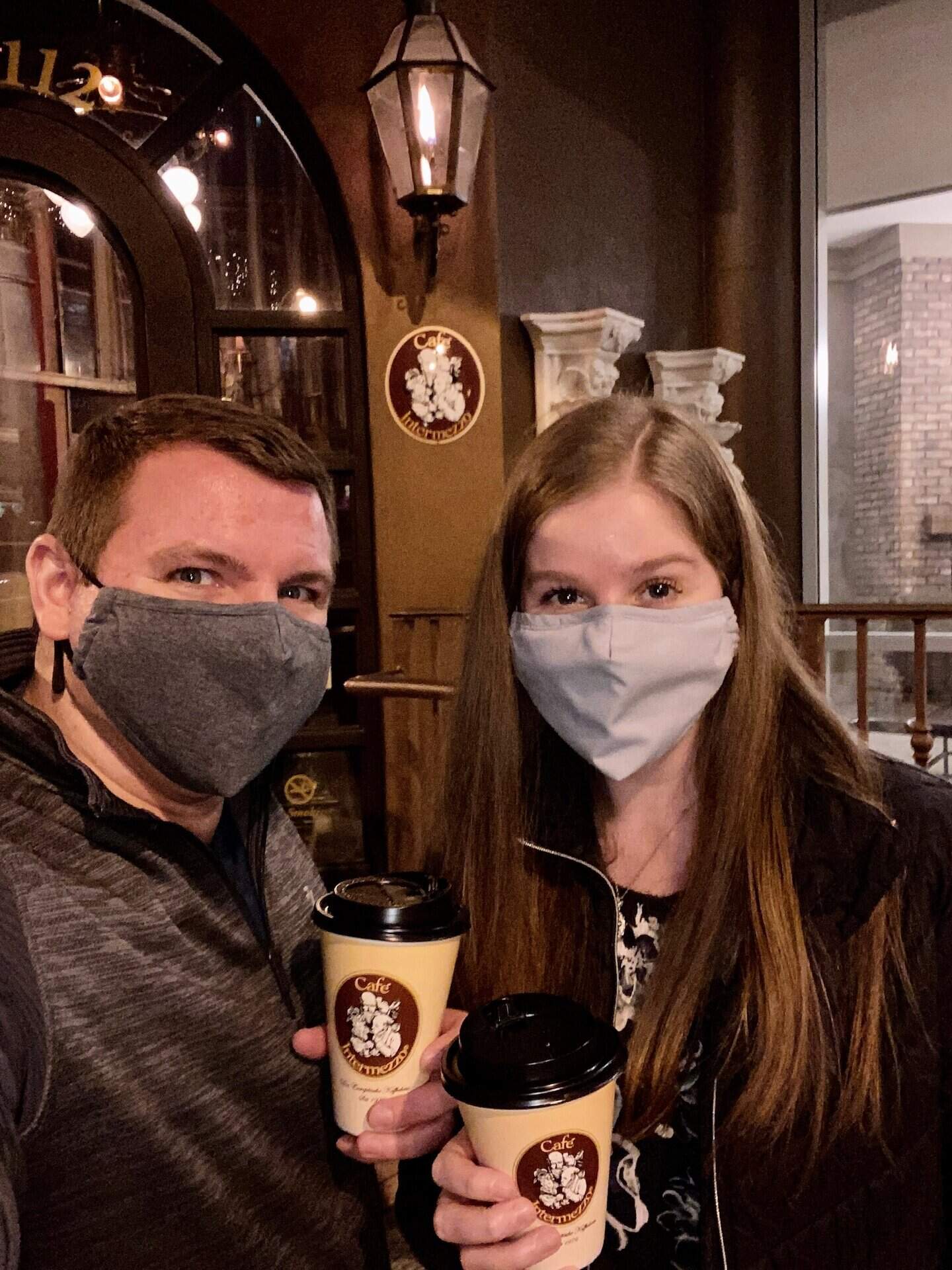 Two people wearing face masks hold coffee cups outside a building at night.