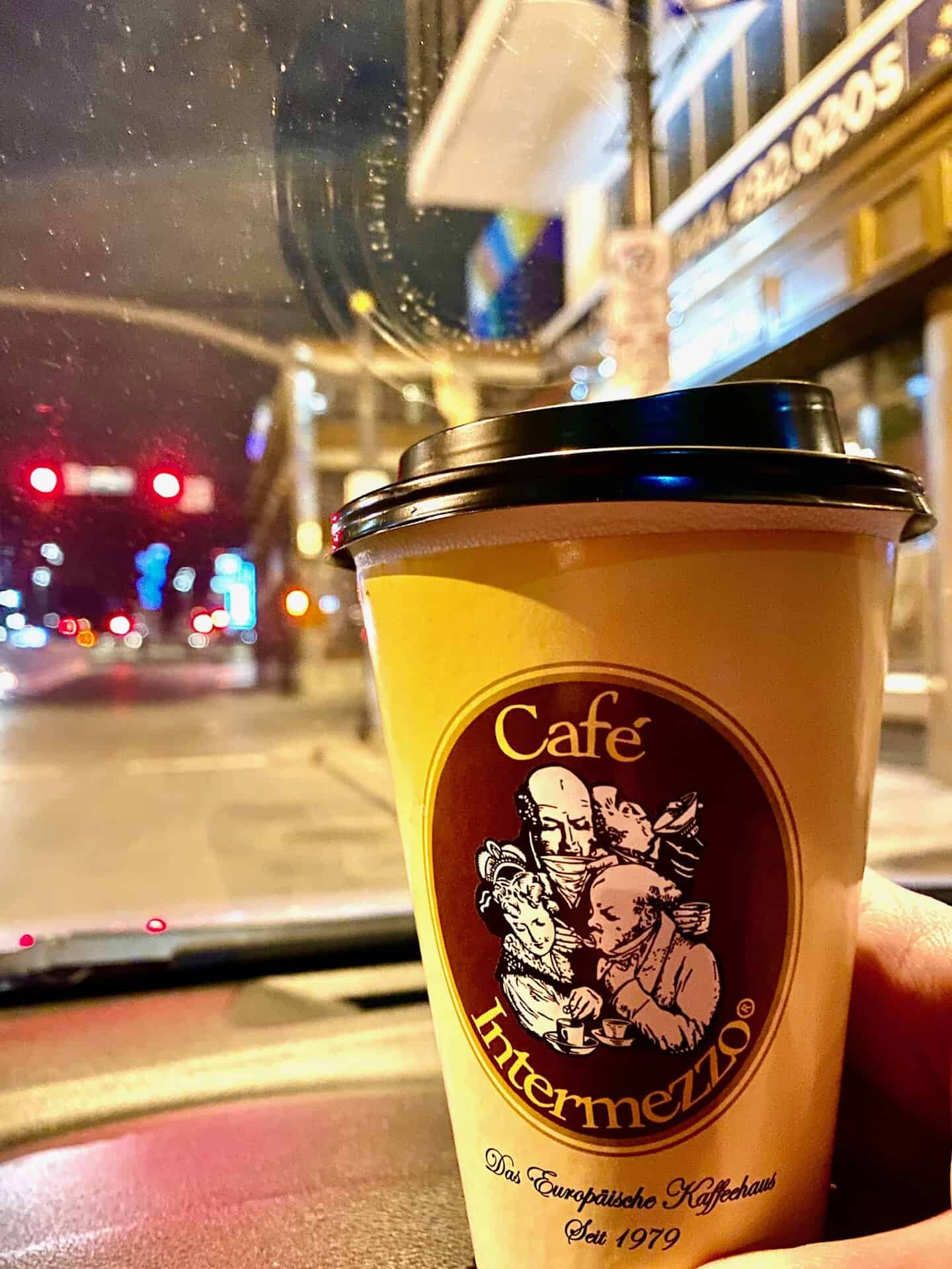 A hand holds a Café Intermezzo coffee cup inside a car at night, with city streetlights and buildings visible through the windshield.
