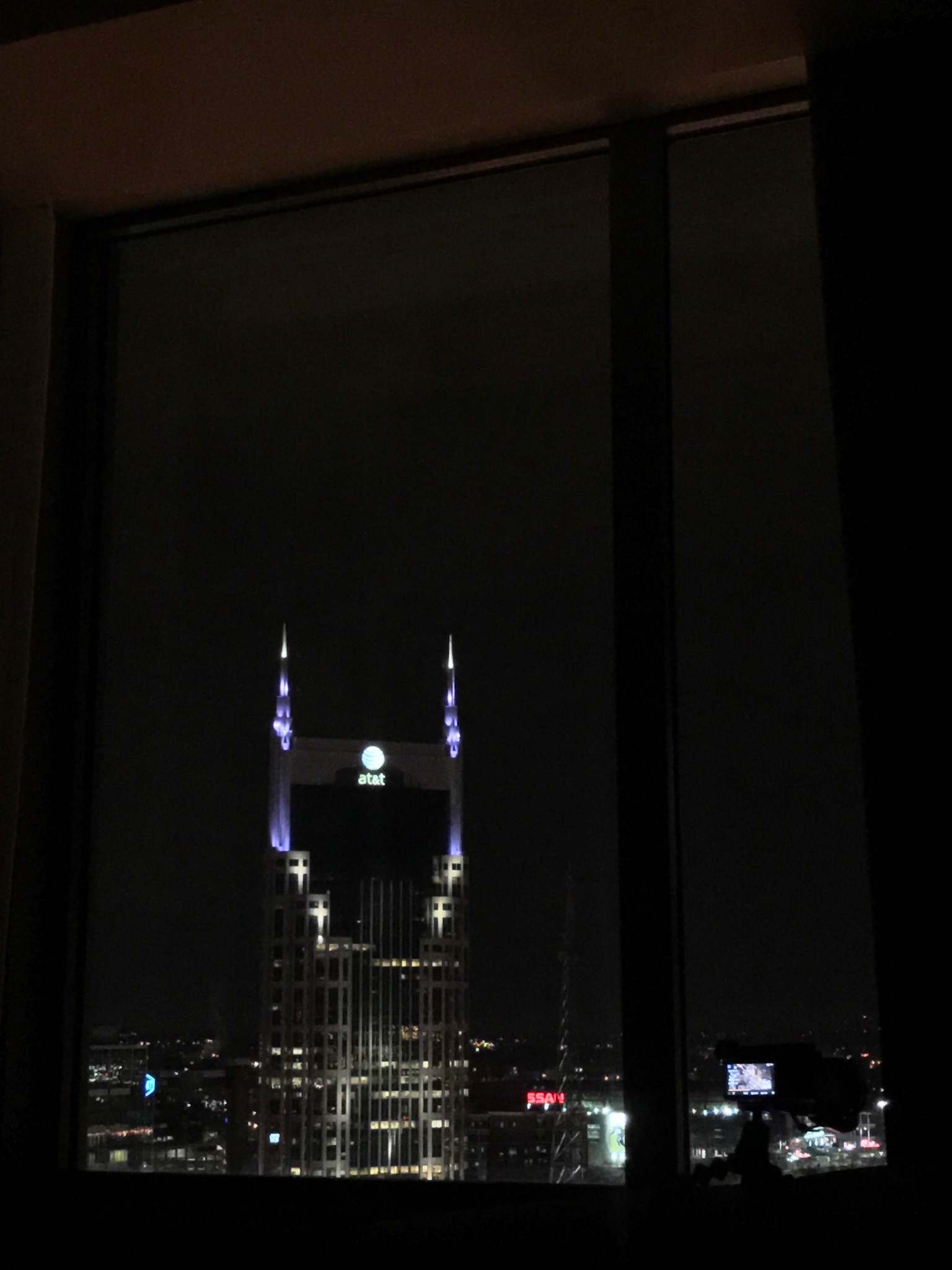 Night view of the AT&T building in Nashville seen through a window, with its spires lit up against the dark sky and a small camera on a tripod visible in the corner.