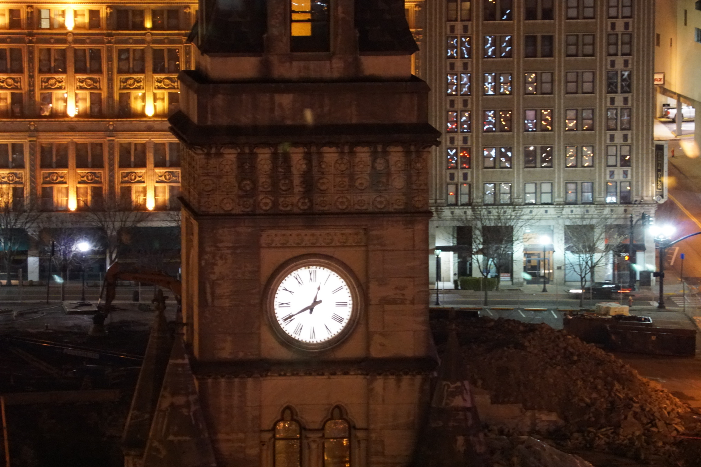 A clock tower displays the time 7:30 in front of illuminated buildings at night, with piles of rubble and construction equipment at its base.