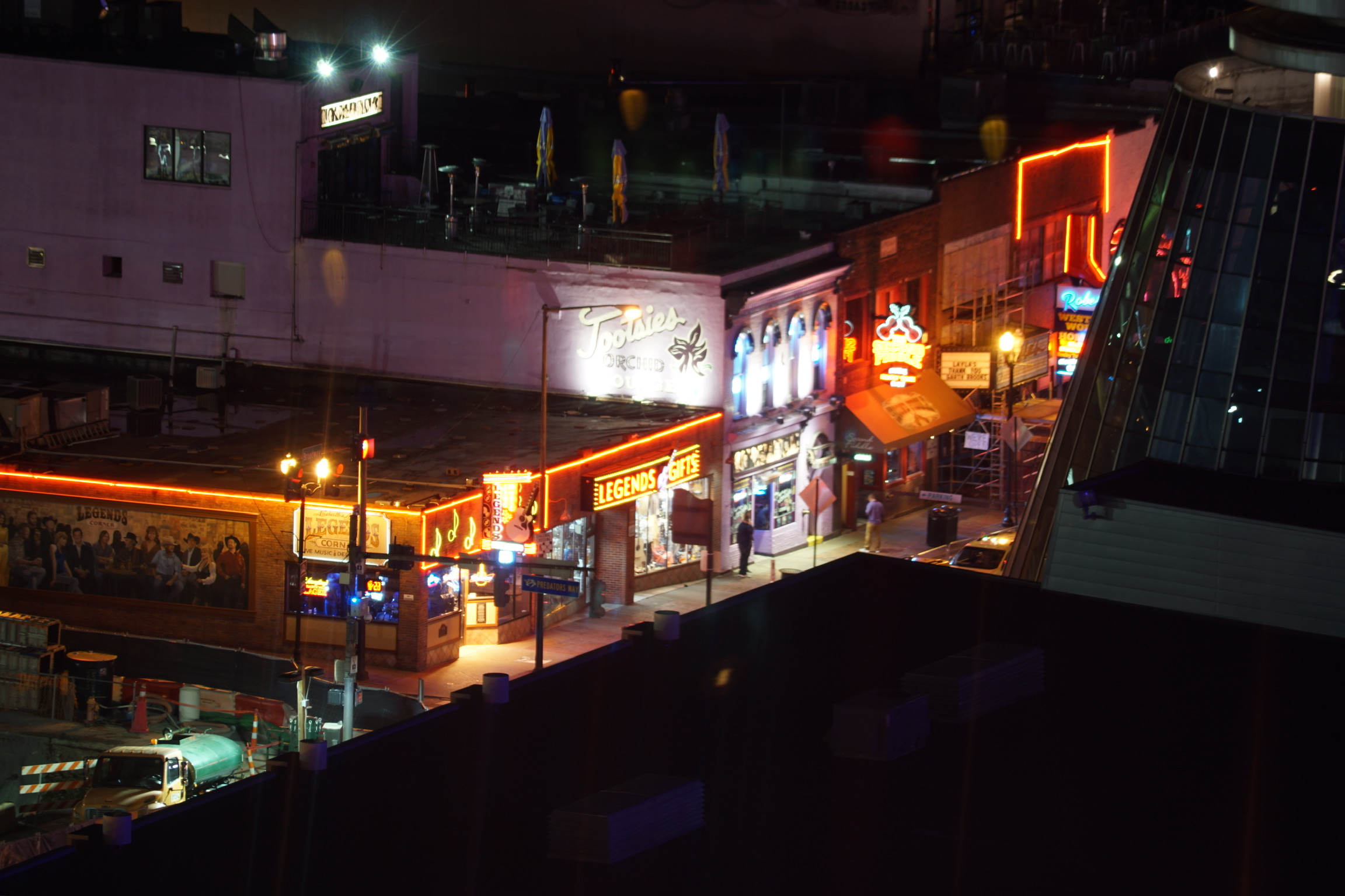 Neon signs illuminate bars and restaurants along a city street at night, with pedestrians walking on the sidewalk.