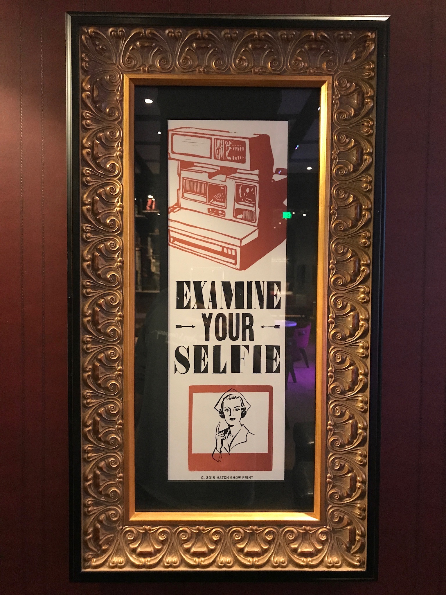 A framed poster with vintage cameras and the text “EXAMINE YOUR SELFIE,” featuring a drawing of a person in the bottom corner.