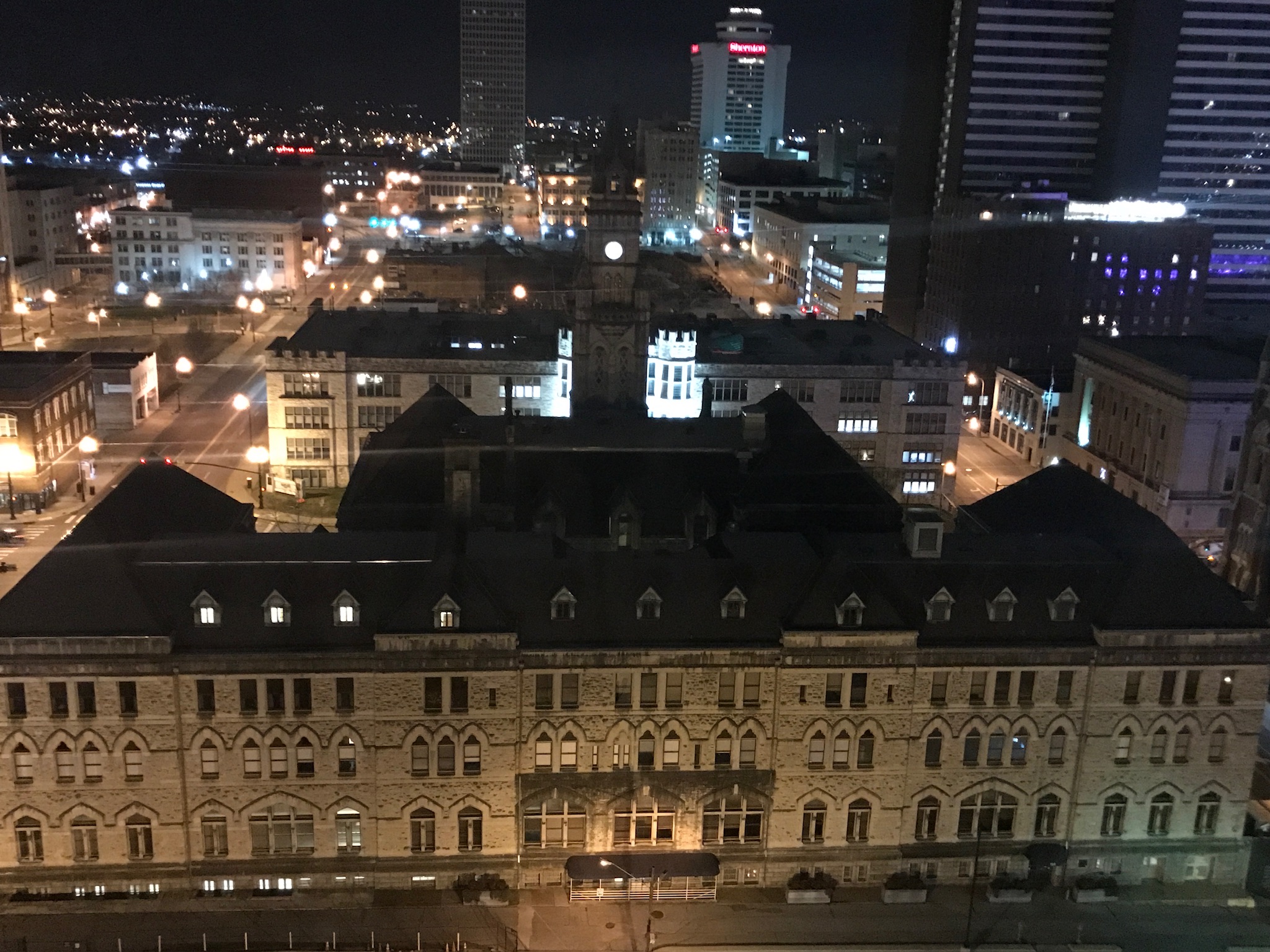 A nighttime cityscape featuring a large historic building in the foreground and modern high-rise buildings illuminated in the background.