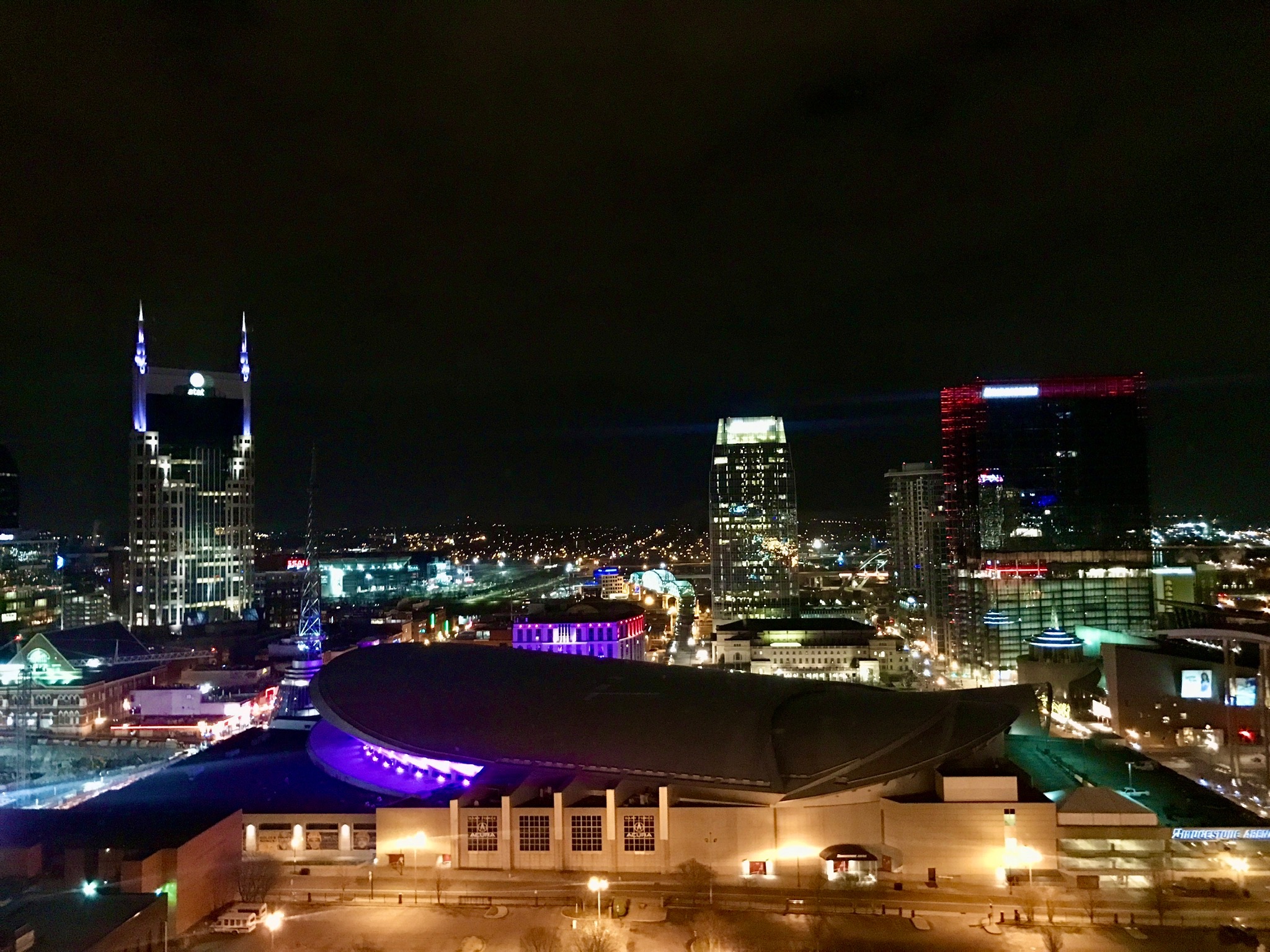 Nighttime cityscape of downtown Nashville featuring illuminated buildings, including the AT&T Building, and the curved roof of the Music City Center in the foreground.