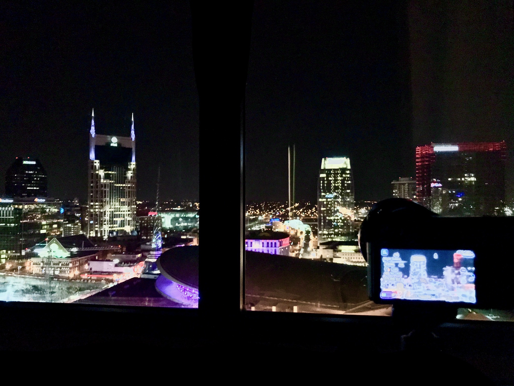 View of a city skyline at night through a window, with illuminated buildings and a camera on a tripod capturing the scene in the foreground.