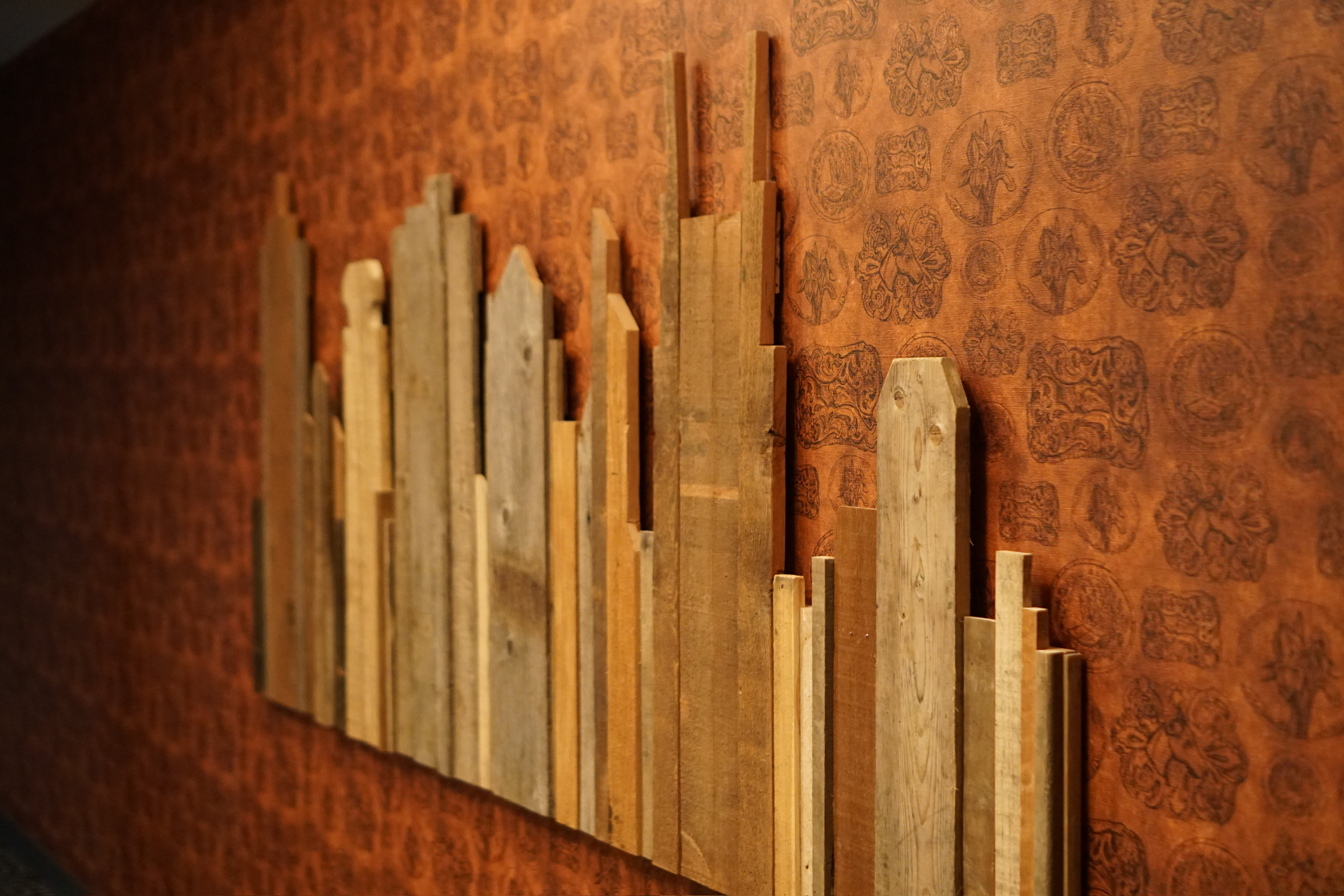 Wooden planks of varying heights are arranged vertically on a patterned orange wall, resembling a city skyline.