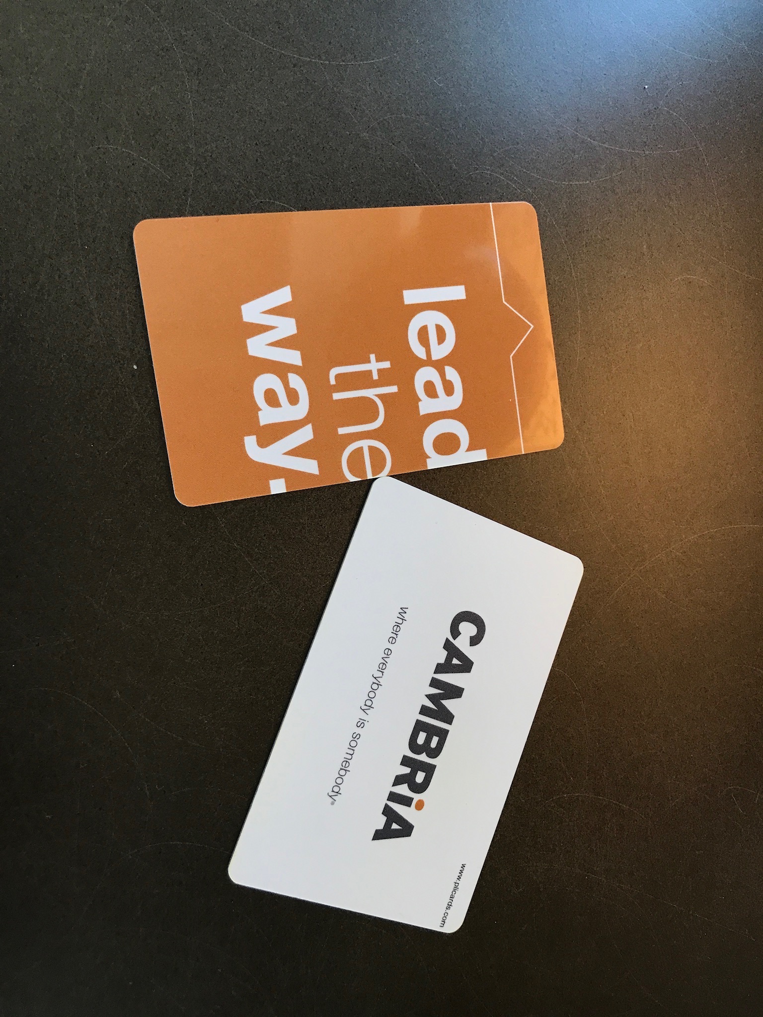 Two hotel key cards on a dark surface; one orange card reads "lead the way," and the other white card displays the "CAMBRIA" hotel logo with a tagline.