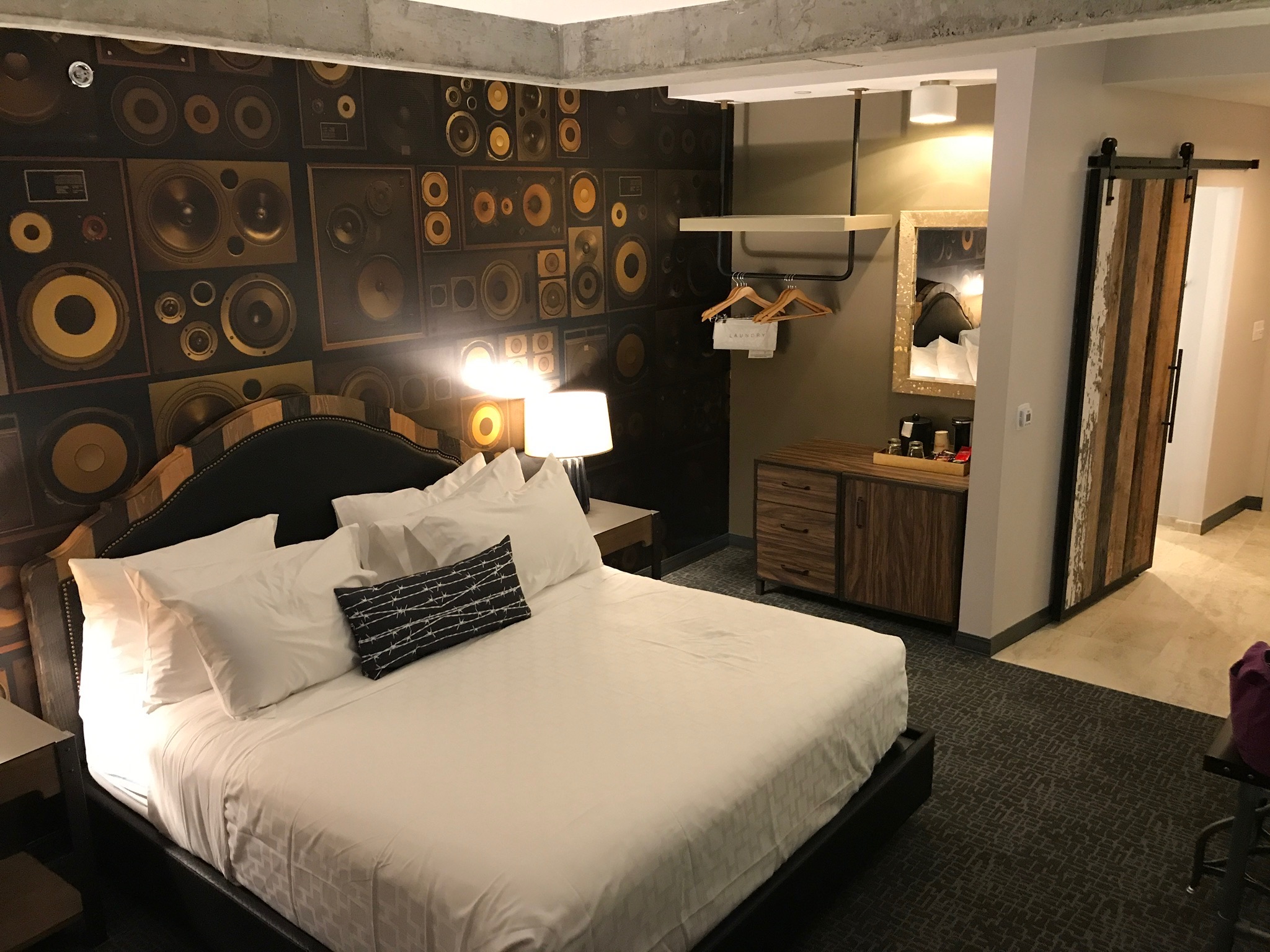 A hotel room with a large bed, speaker-themed accent wall, lamp, open closet with hangers, wooden dresser, and a sliding barn door leading to the bathroom.