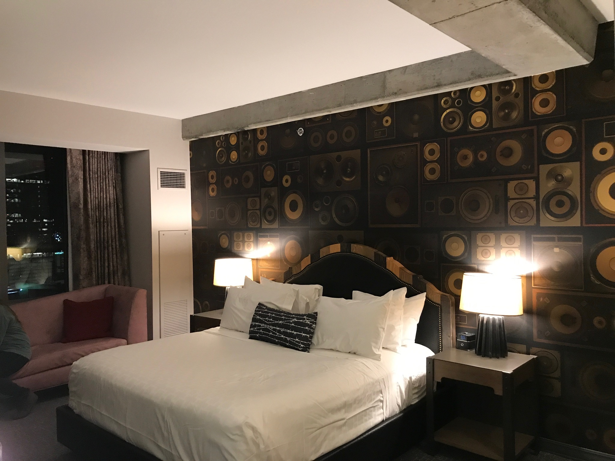 A modern hotel room featuring a bed with white linens, two bedside tables with lamps, and a wall decorated with a speaker-themed mural.