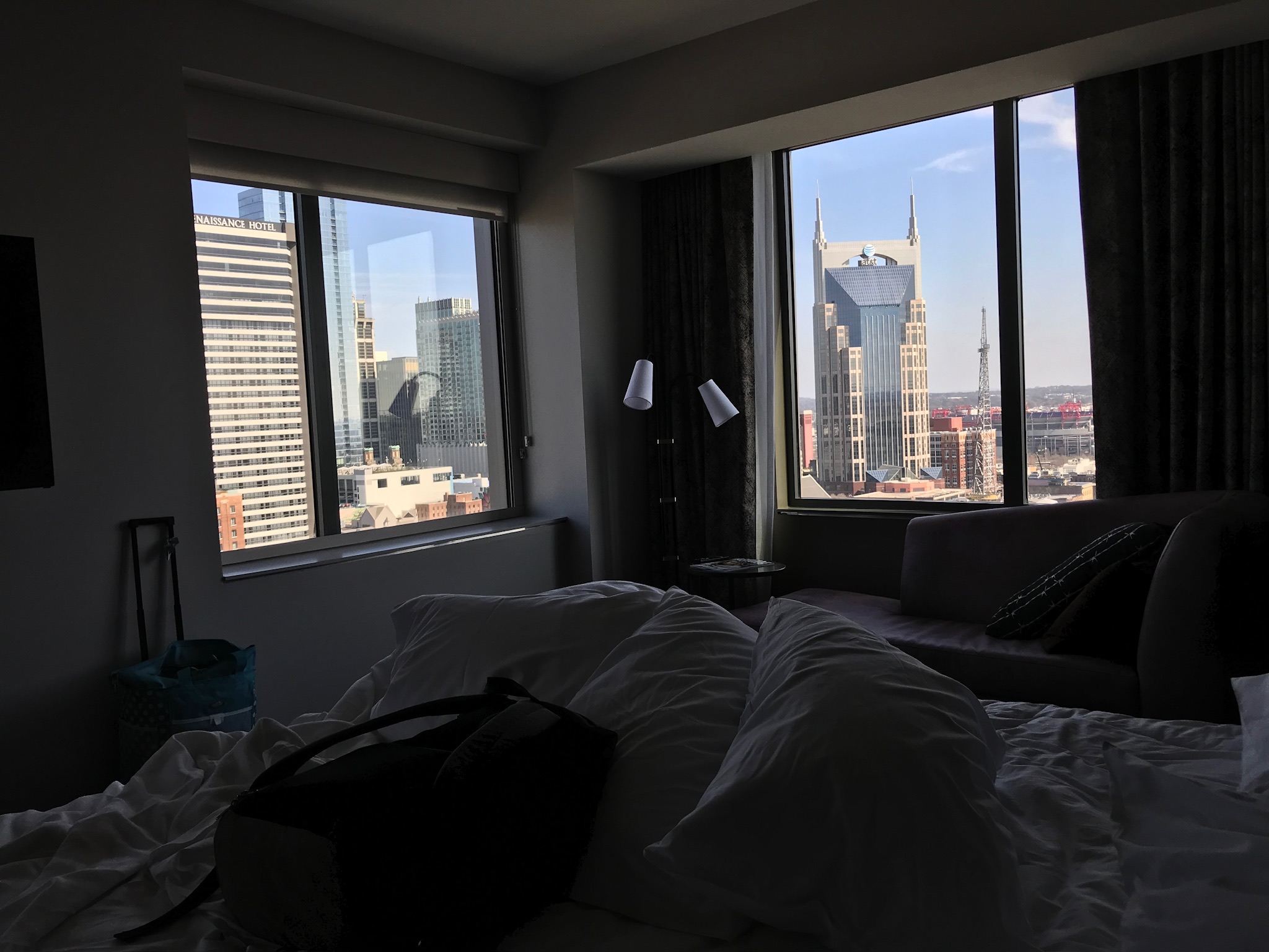 A hotel room with unmade bed and city view through large windows, including modern skyscrapers and the AT&T building in the background.