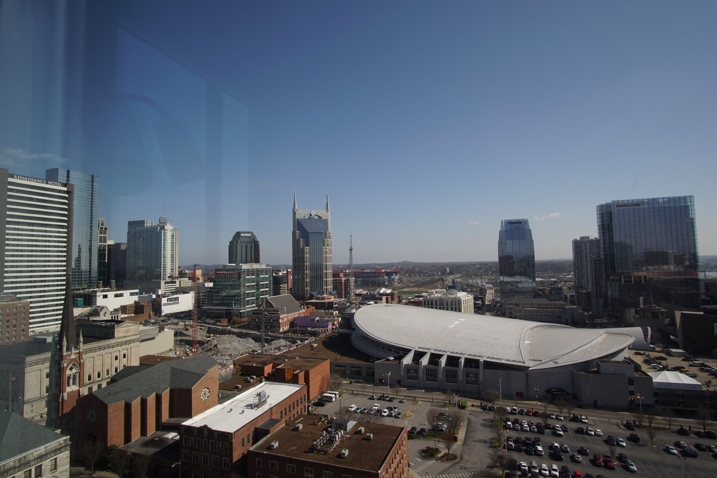 A cityscape view of downtown Nashville, featuring modern skyscrapers, the AT&T Building, and the white-roofed Bridgestone Arena under a clear blue sky.