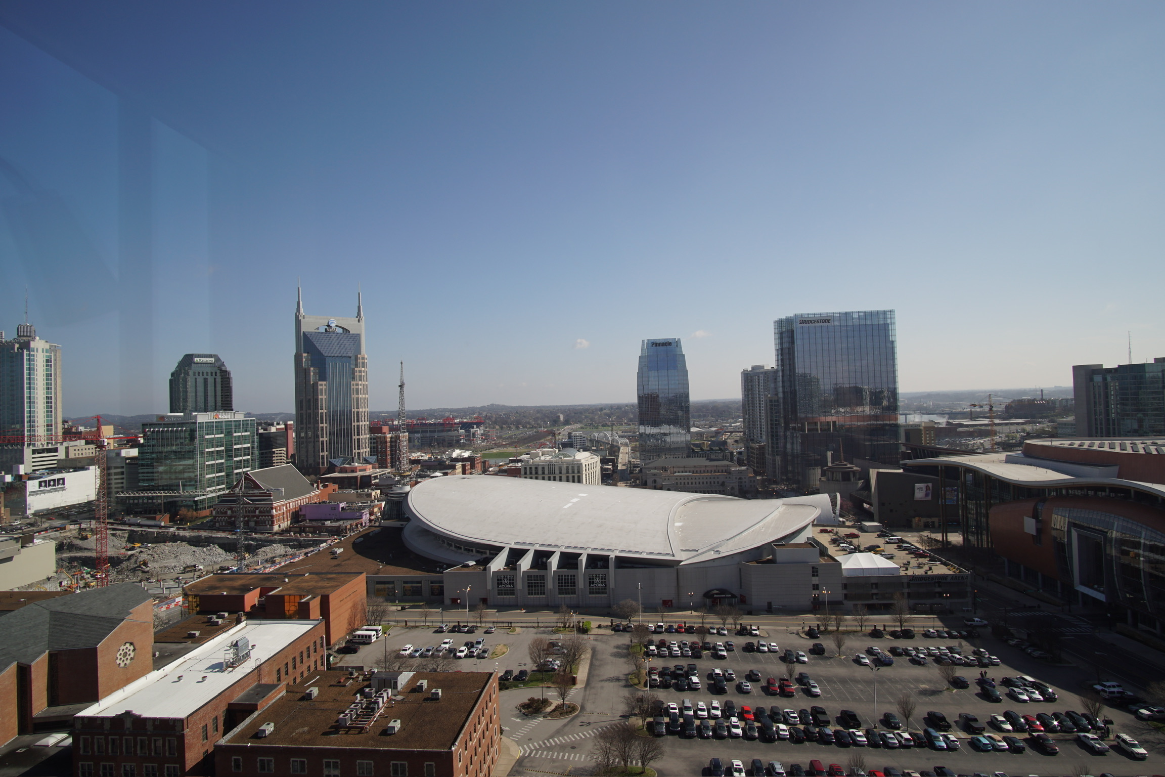 Aerial view of downtown Nashville, featuring the distinctive AT&T Building, Bridgestone Arena with its white roof, and surrounding high-rise buildings under a clear blue sky.