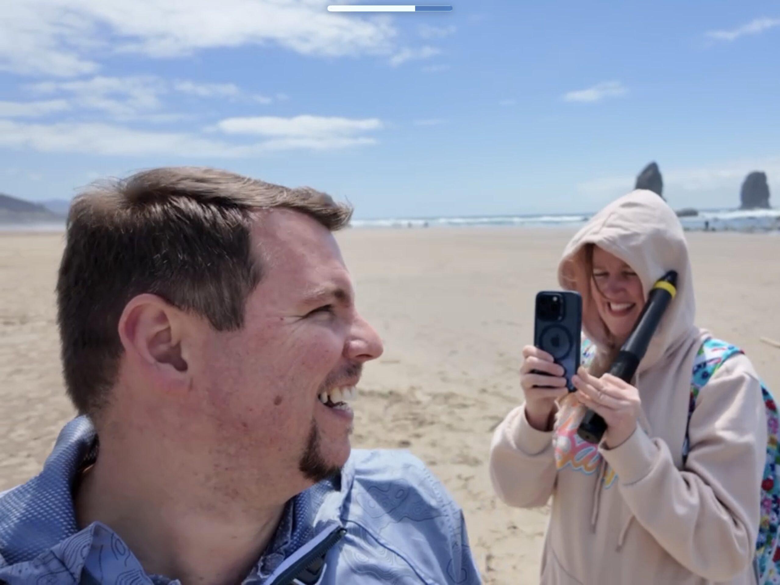 Two people stand on a sandy beach; one smiles at the camera while the other, wearing a hood, holds a smartphone and a small device, with rocks and the ocean in the background.