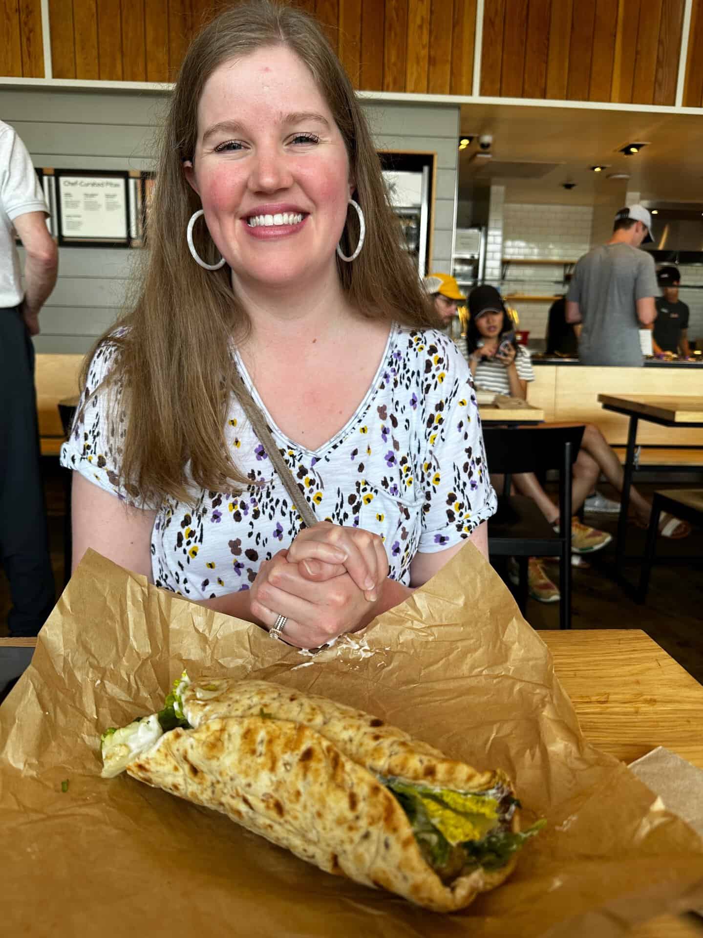 A person beams at a table inside a casual dining spot, eagerly anticipating the flavors of a large wrapped Nashville-style hot chicken sandwich resting on brown paper before them.