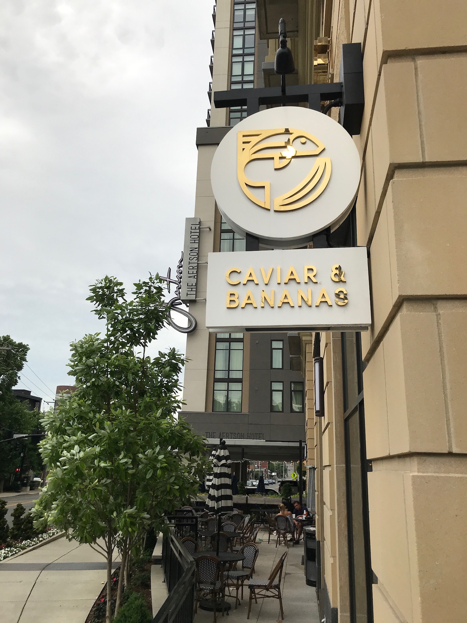 Sign for "Caviar & Bananas" hangs on a beige building above an outdoor dining area with tables, chairs, and umbrellas; trees and other buildings are visible nearby.