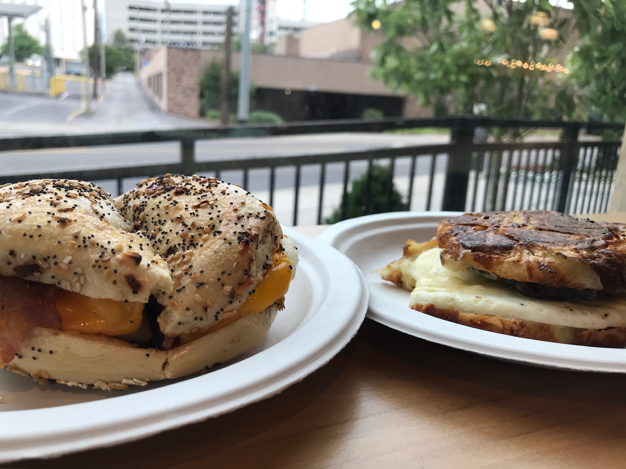 Two breakfast sandwiches on paper plates sit on a wooden table by a window, showing a view of buildings and trees outside.