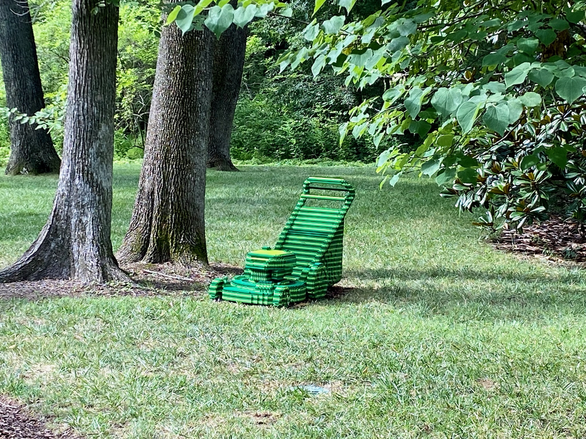A green sculpture resembling a lawnmower, made from what appears to be stacked rectangular objects, sits on grass near several trees.