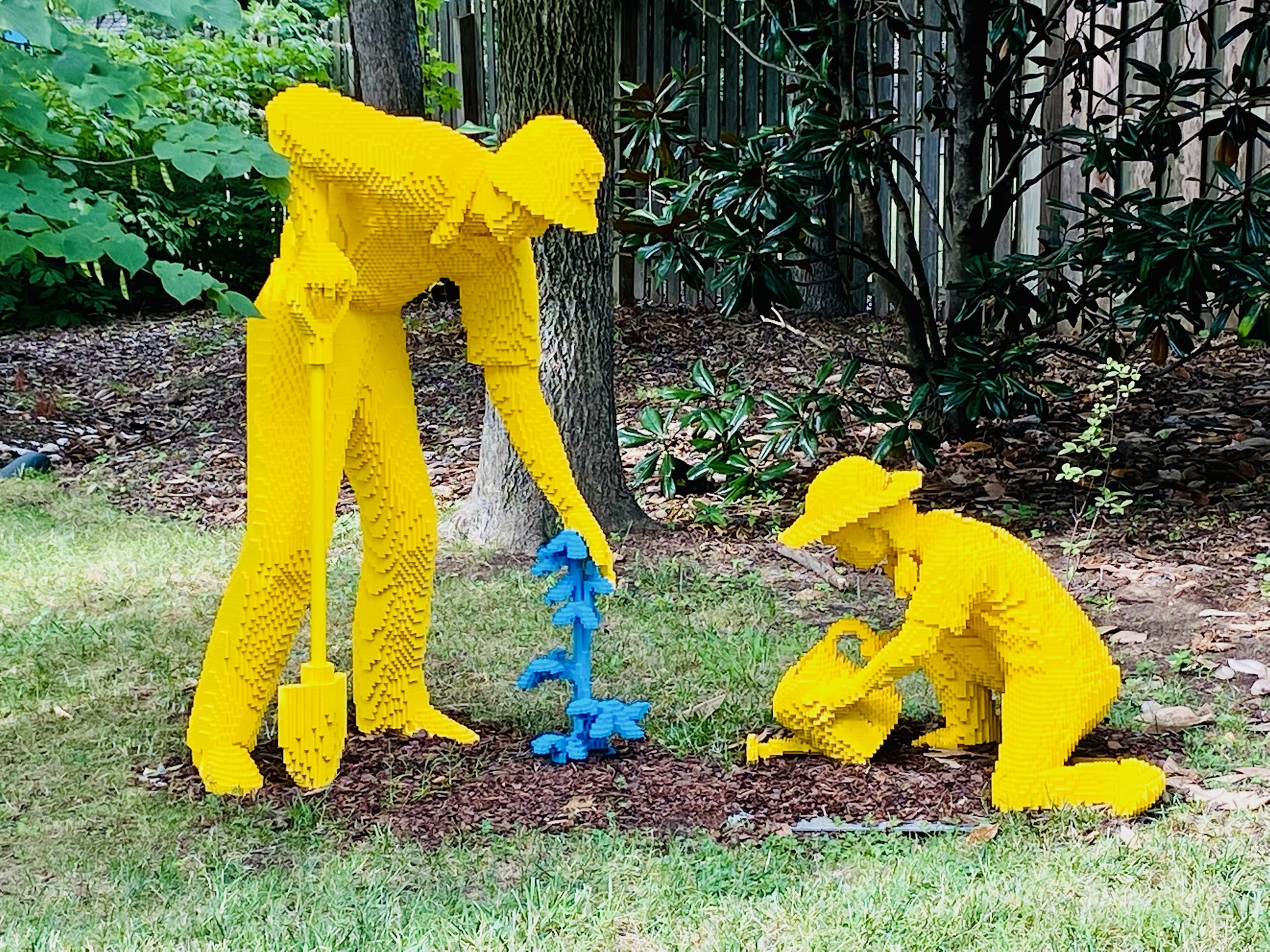 Two yellow Lego sculptures depict a standing adult with a shovel and a kneeling child watering a blue plant in a garden setting.
