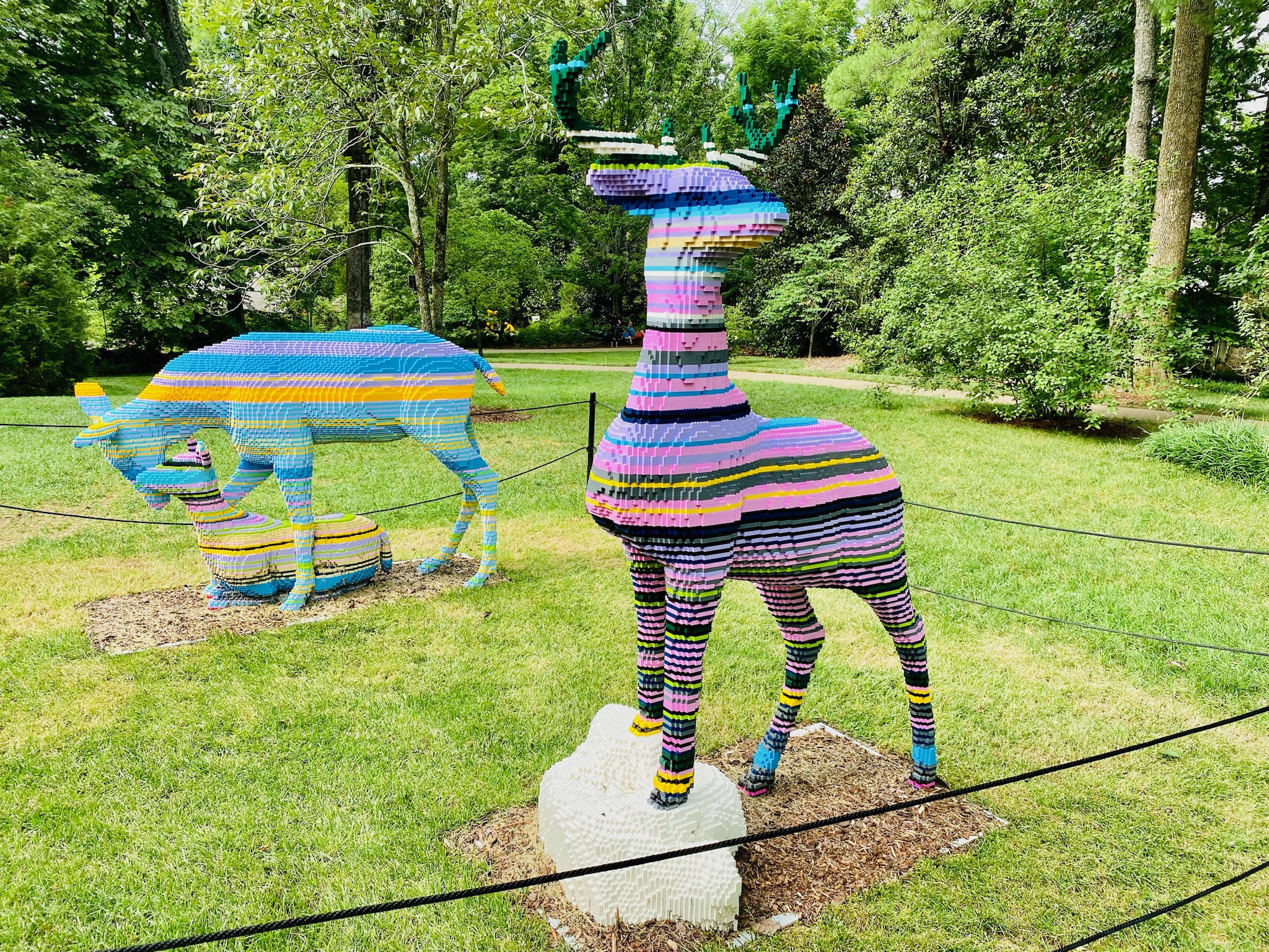 Two deer sculptures made of colorful stacked bricks are displayed outdoors on grass, surrounded by trees and rope barriers.