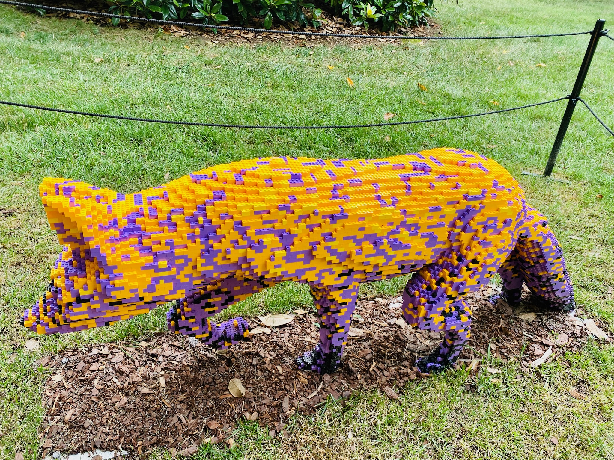 A life-size fox sculpture made from yellow, orange, and purple LEGO bricks stands on mulch in a grassy outdoor area.