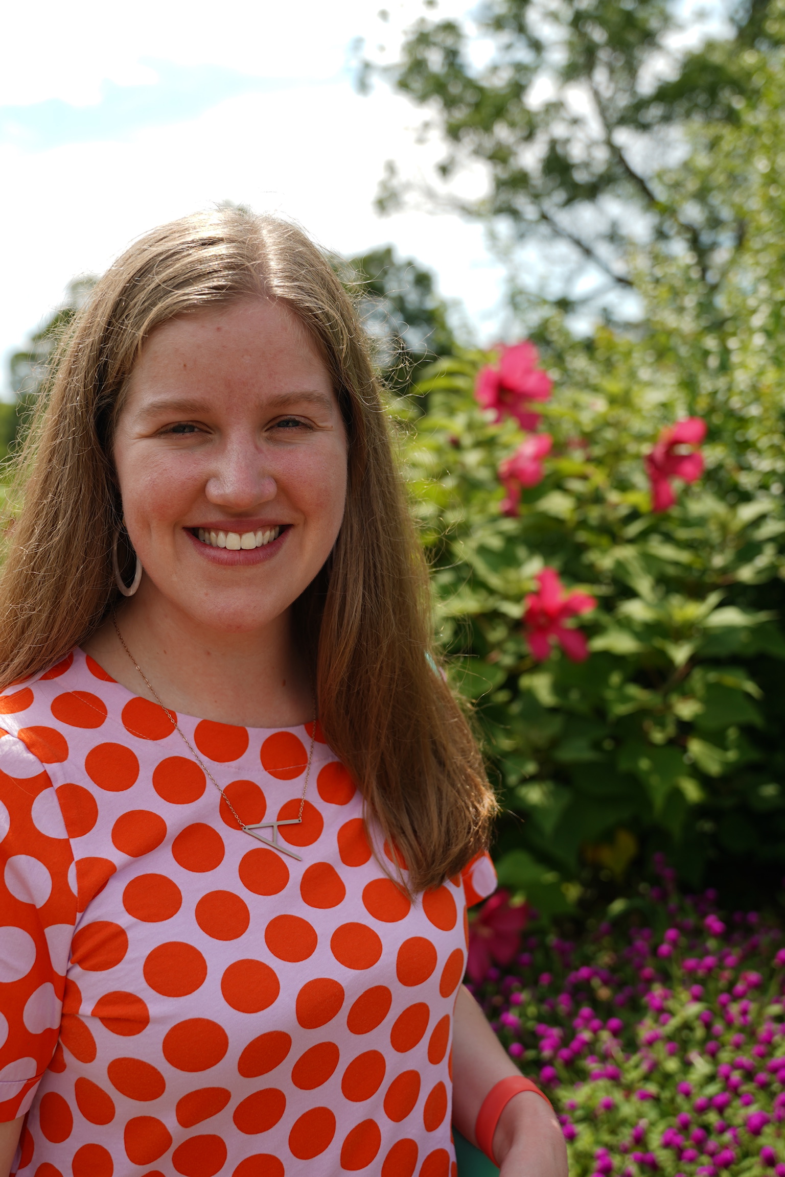 A woman with long, light brown hair wearing an orange and white polka dot shirt stands outdoors in front of blooming flowers and greenery, smiling at the camera.