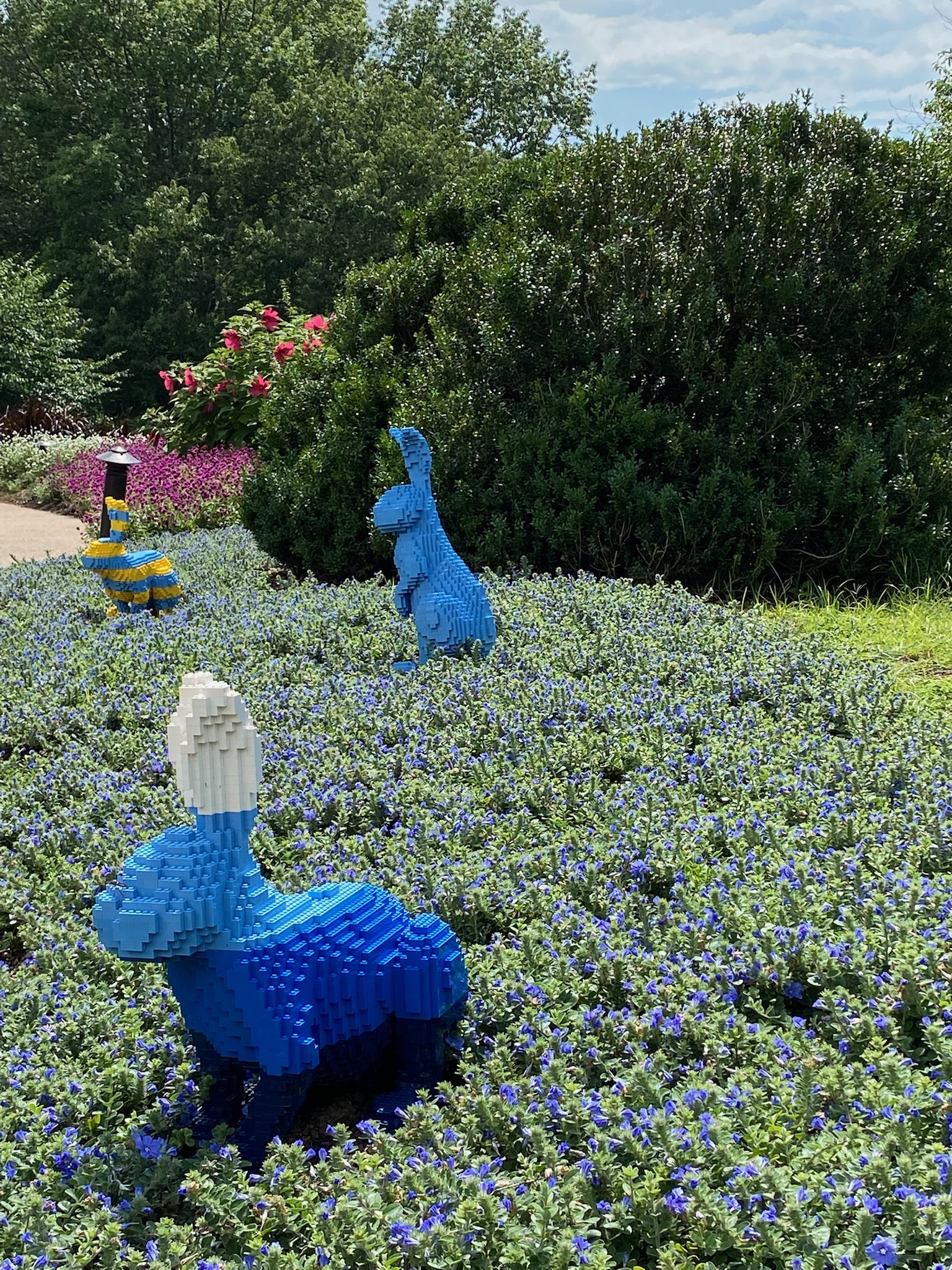 Three blue LEGO bunny sculptures are placed among purple flowers and greenery in an outdoor garden setting on a sunny day.