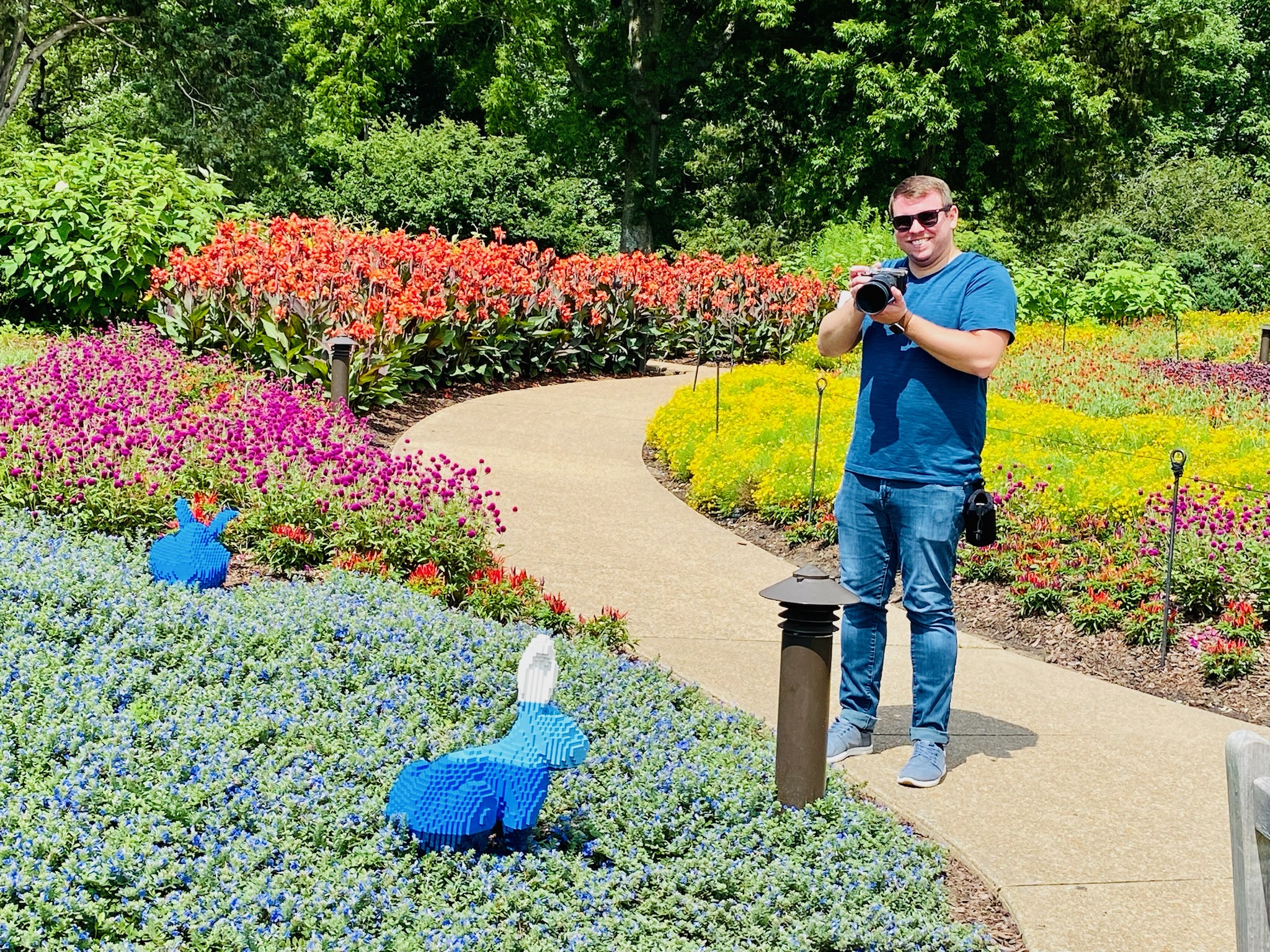A man with a camera stands on a garden path surrounded by colorful flowers and large blue LEGO bunny sculptures.