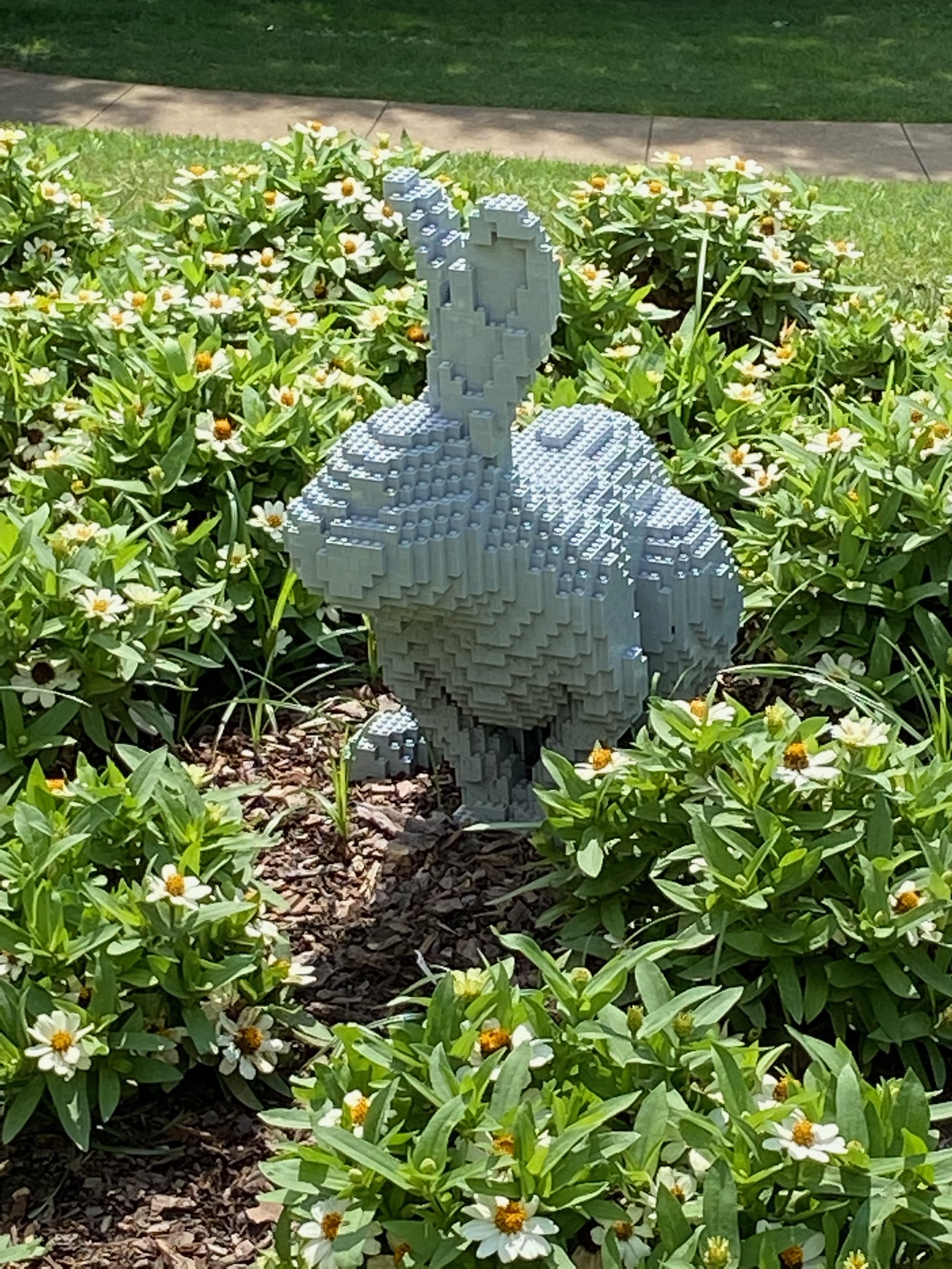 A rabbit sculpture made of stacked gray LEGO bricks is displayed among green plants and white flowers in an outdoor garden.