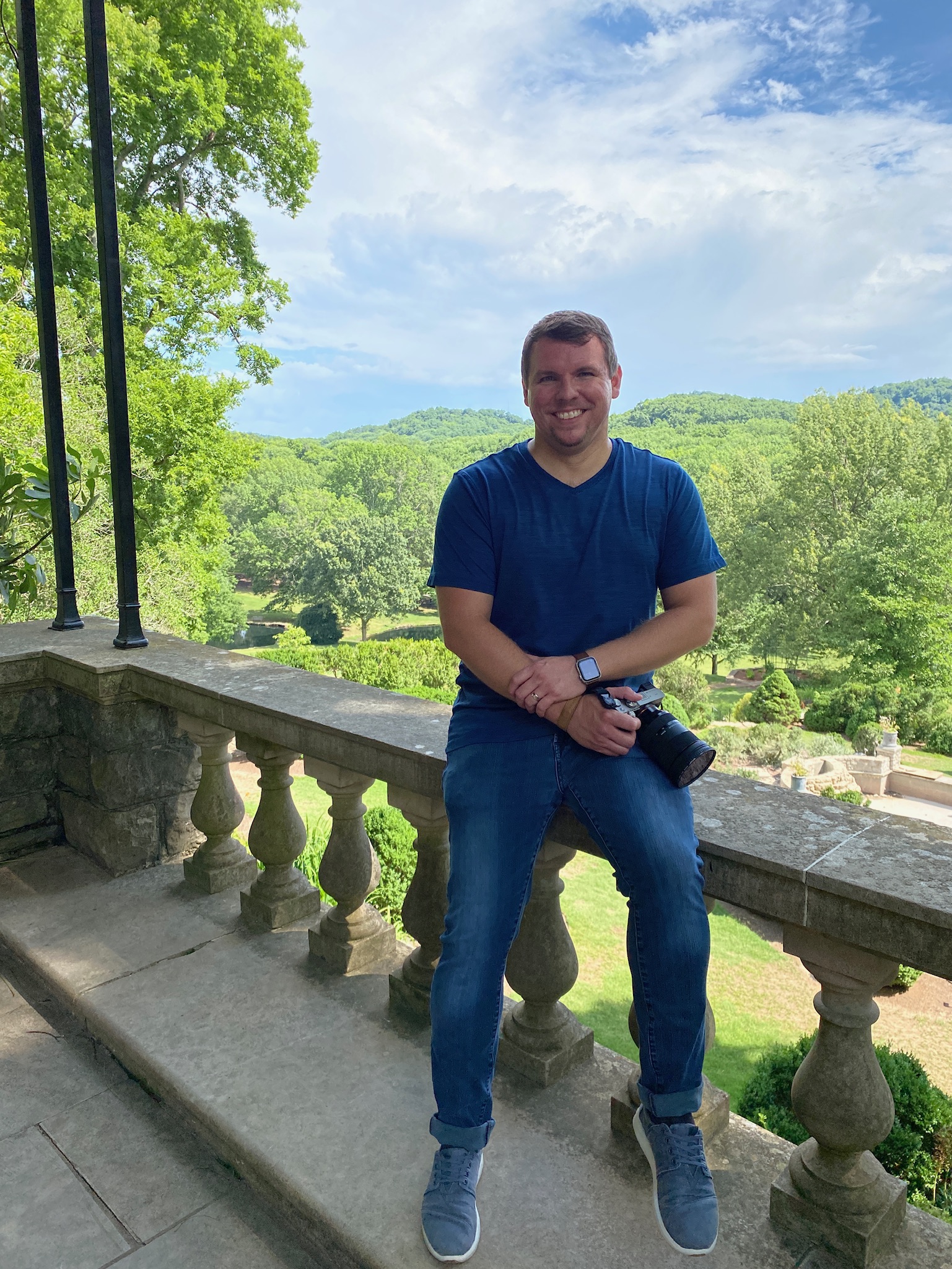 A man in a blue t-shirt and jeans sits on a stone railing holding a camera, with a scenic green landscape and partly cloudy sky in the background.