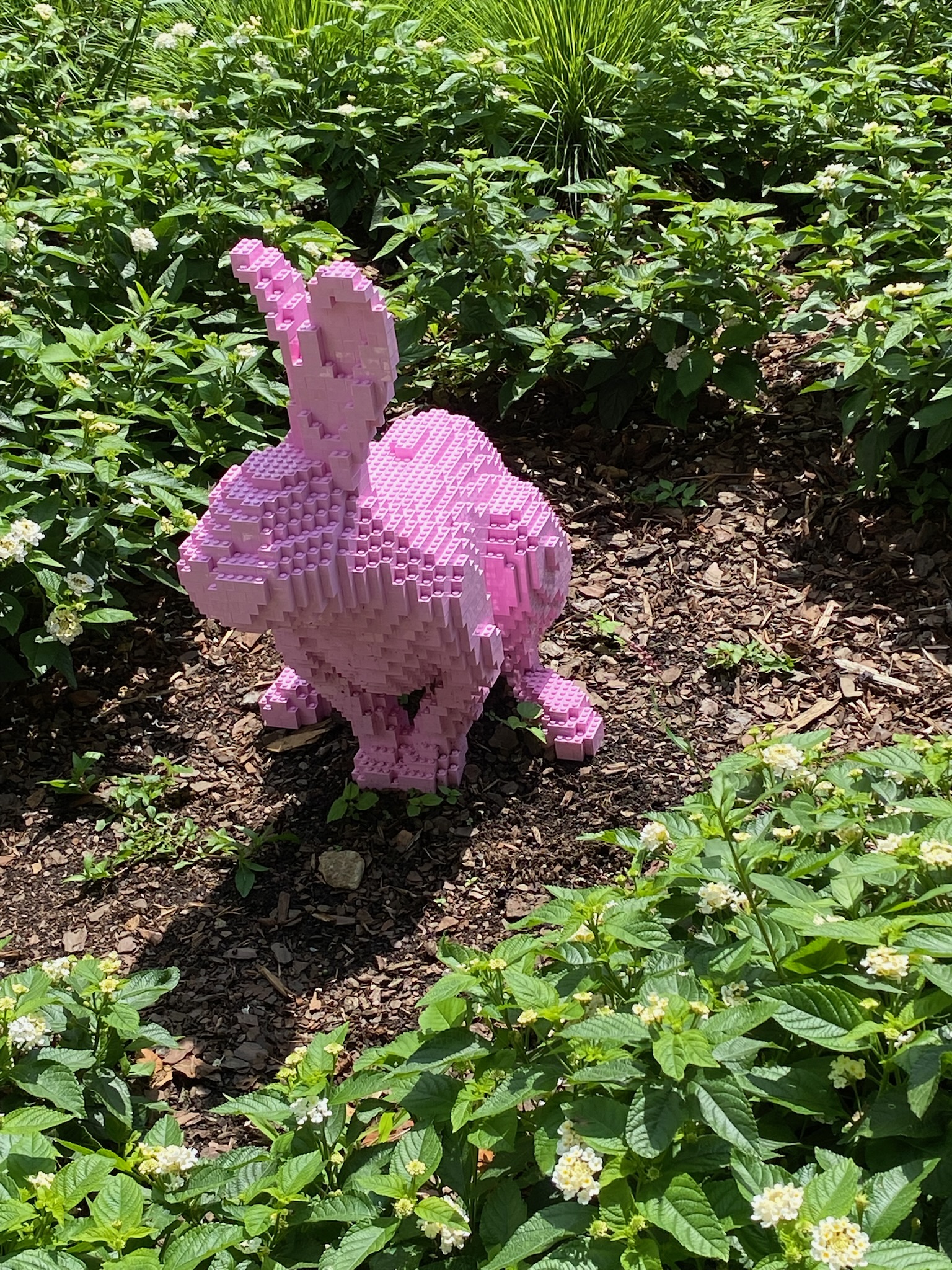 A pink rabbit sculpture made of LEGO bricks sits on a patch of soil surrounded by green plants and small white flowers.