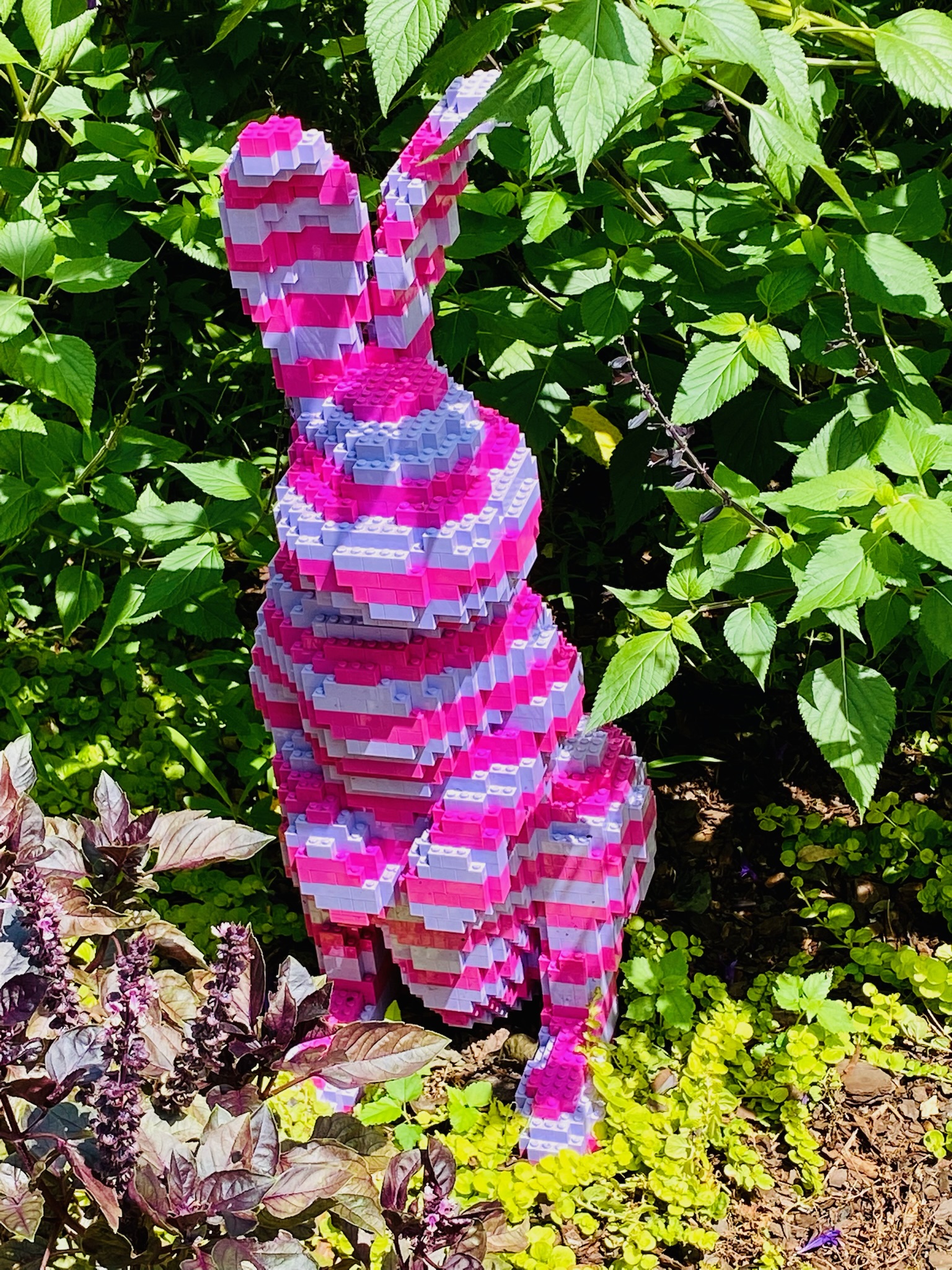 A sculpture of a rabbit made from pink, purple, and white LEGO bricks is placed among green plants outdoors.