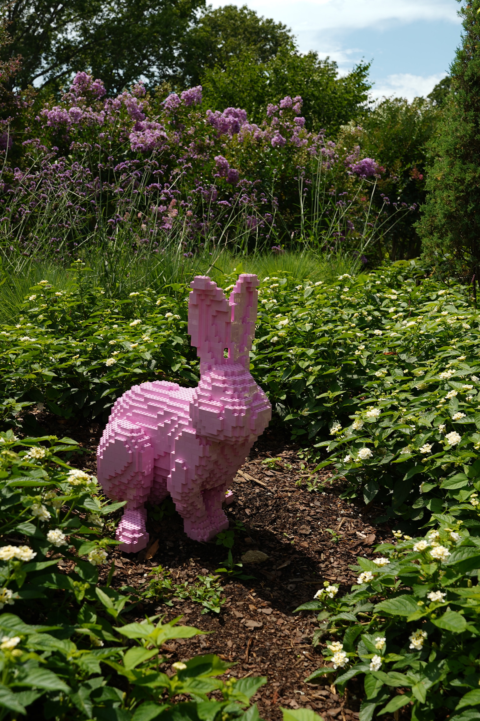 A pink rabbit sculpture made of LEGO bricks stands among green plants and white flowers in a garden with purple-flowered bushes in the background.