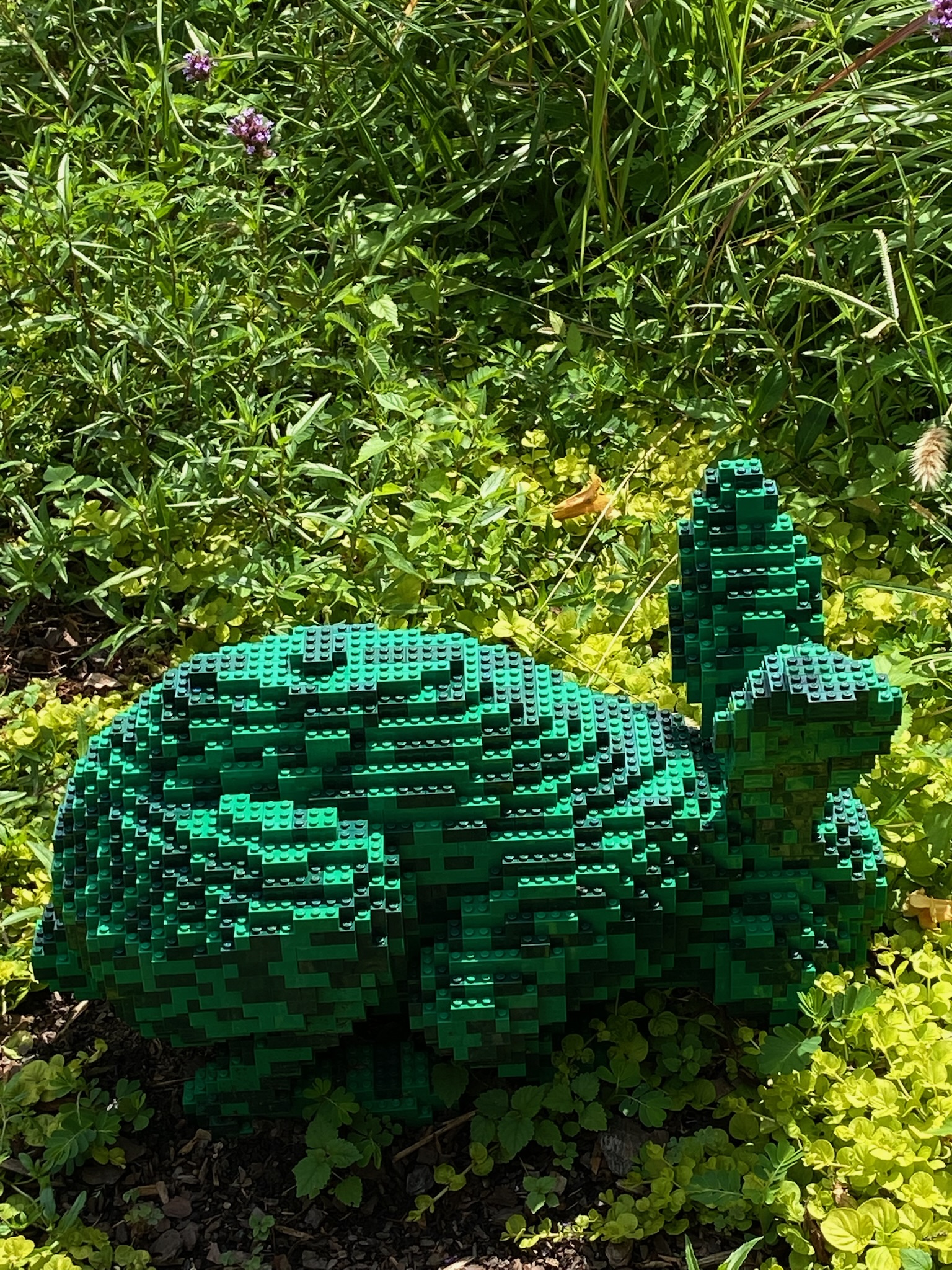 A green rabbit sculpture made of LEGO bricks sits on the ground amid green plants and grass, outdoors in bright sunlight.