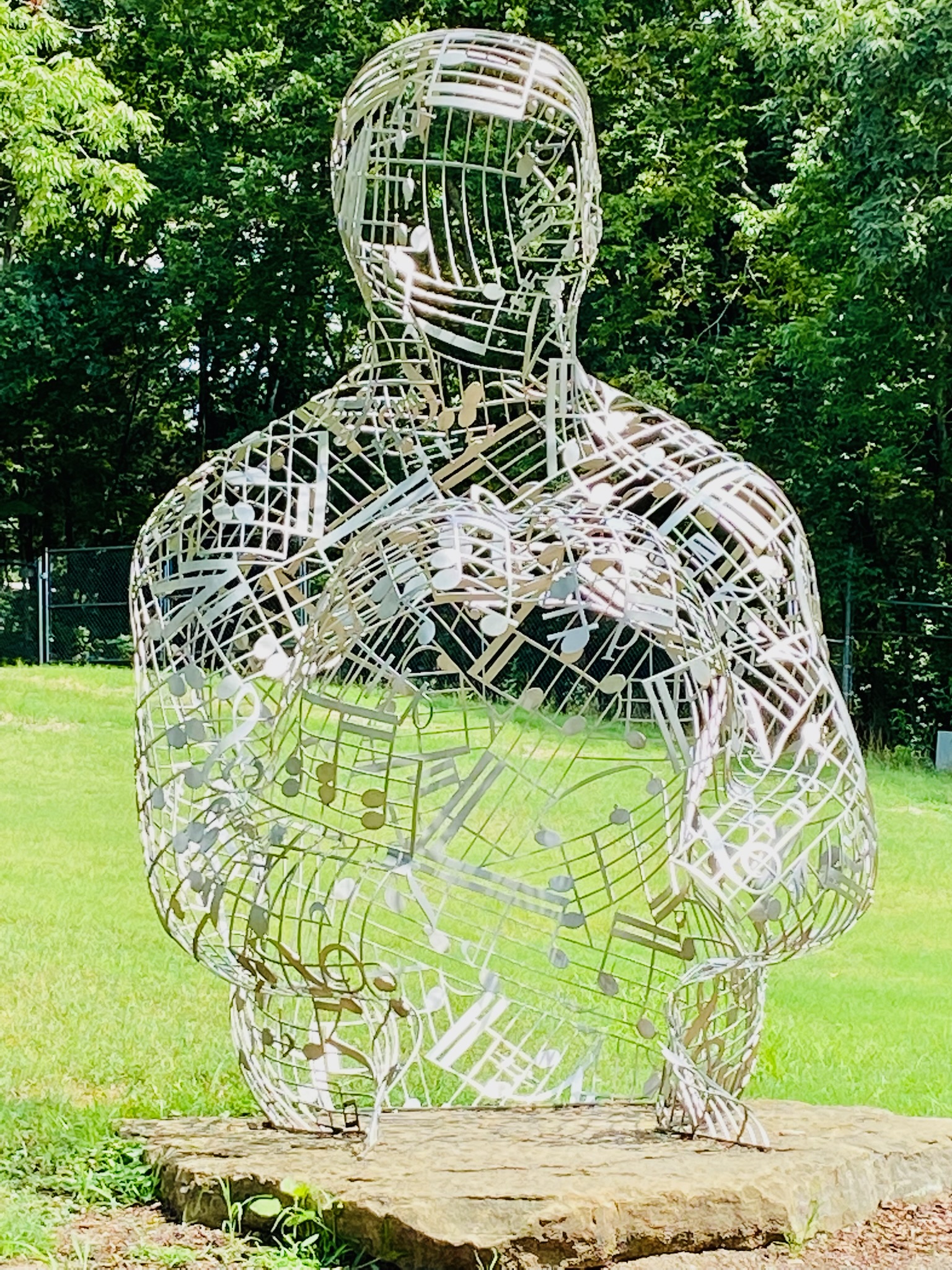 A metal sculpture of a human figure composed of musical staffs and notes stands on a stone base in a grassy outdoor setting with trees in the background.