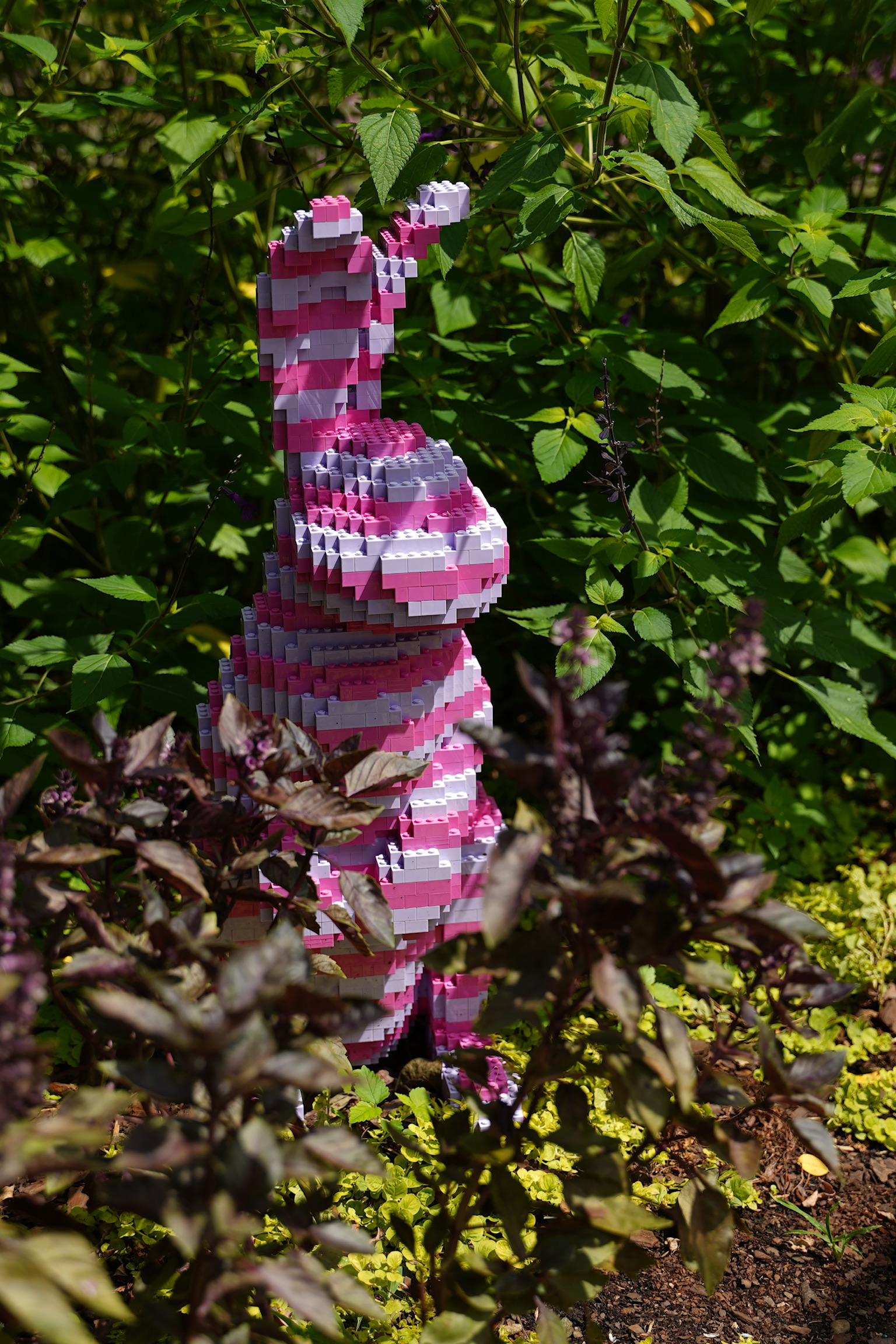 A pink and white rabbit sculpture made of stacked LEGO bricks stands among green plants and foliage in a garden.