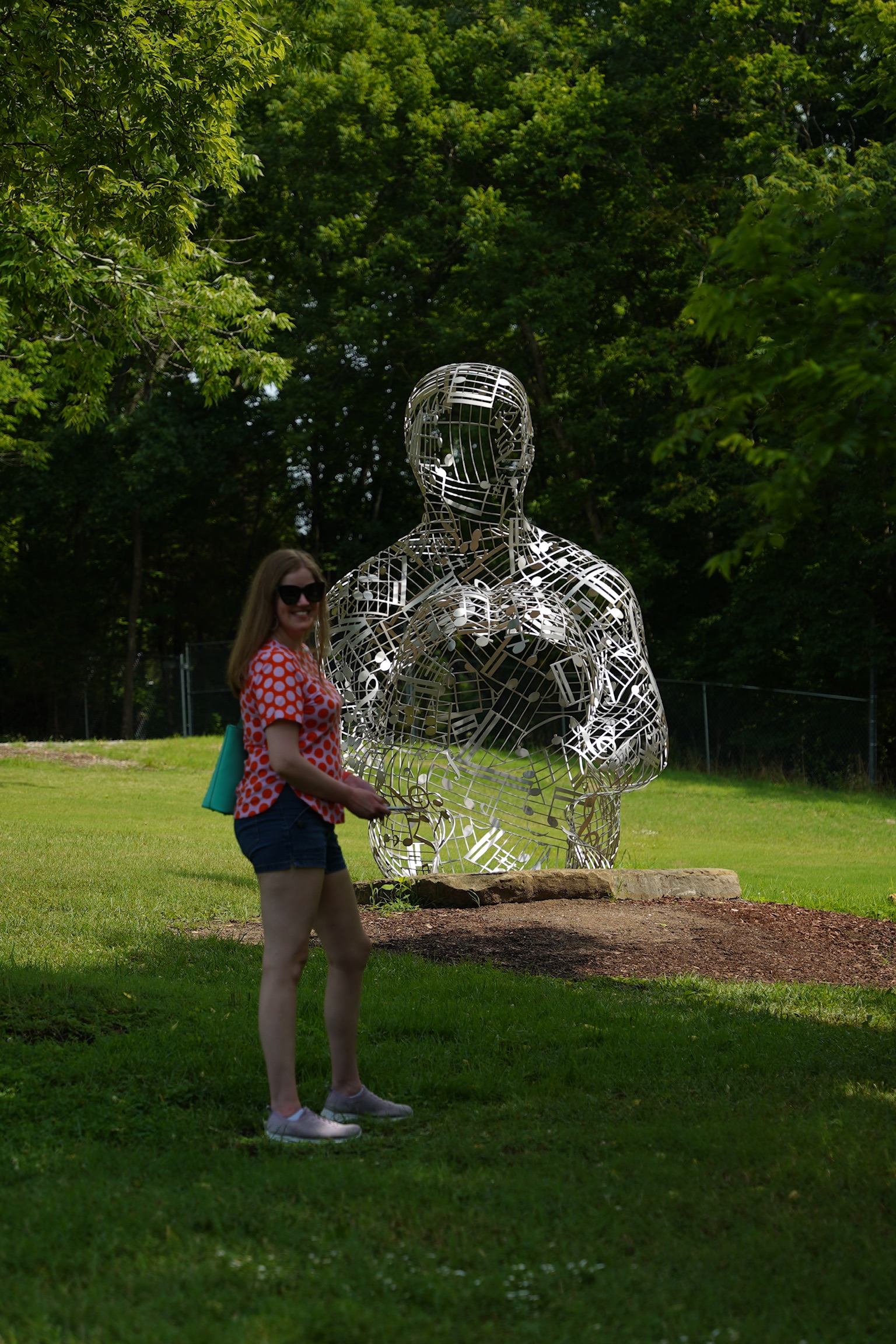 A woman stands on grass near a large metal wire sculpture of a seated human figure, with trees and a fence in the background.