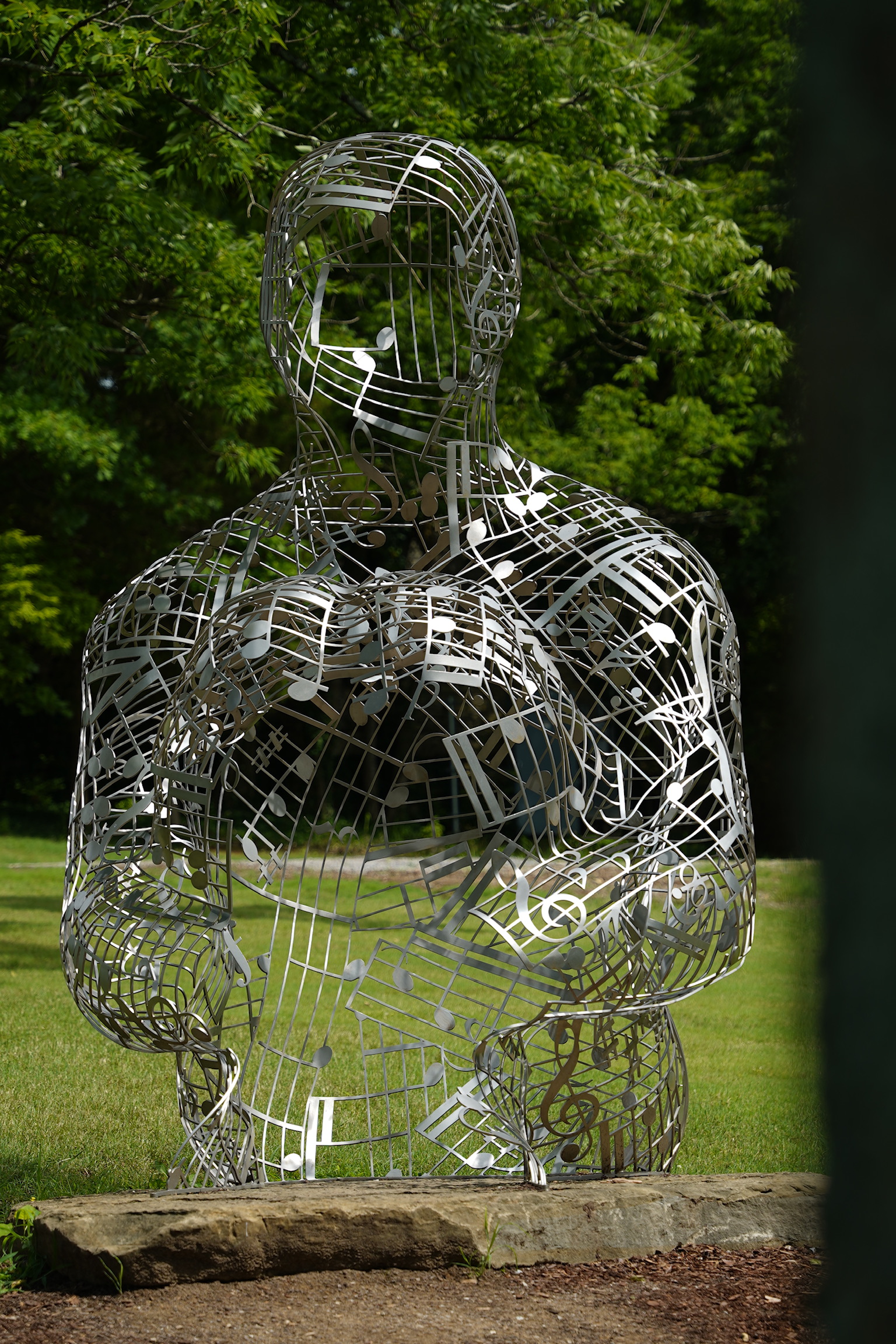 A metal sculpture of a human figure made from interconnected musical notes and symbols stands on grass with trees in the background.