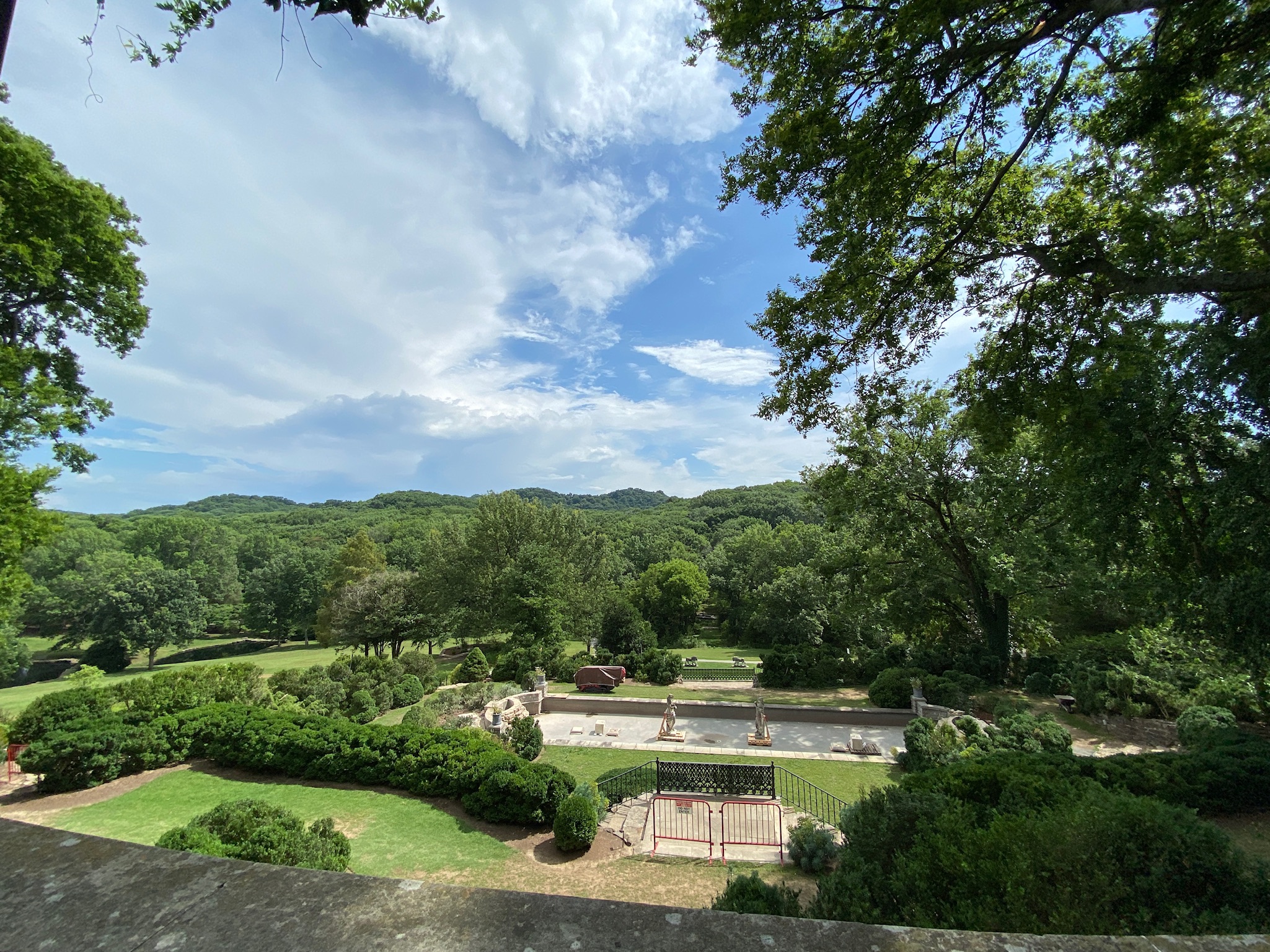 A landscaped garden with trees, shrubs, a paved area, and benches, set against a backdrop of green hills under a partly cloudy sky.