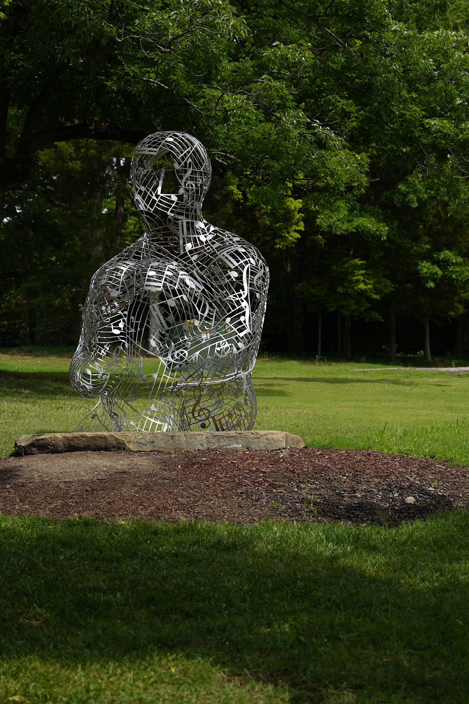 A metal wire sculpture of a seated human figure is displayed on a grassy area with trees in the background.