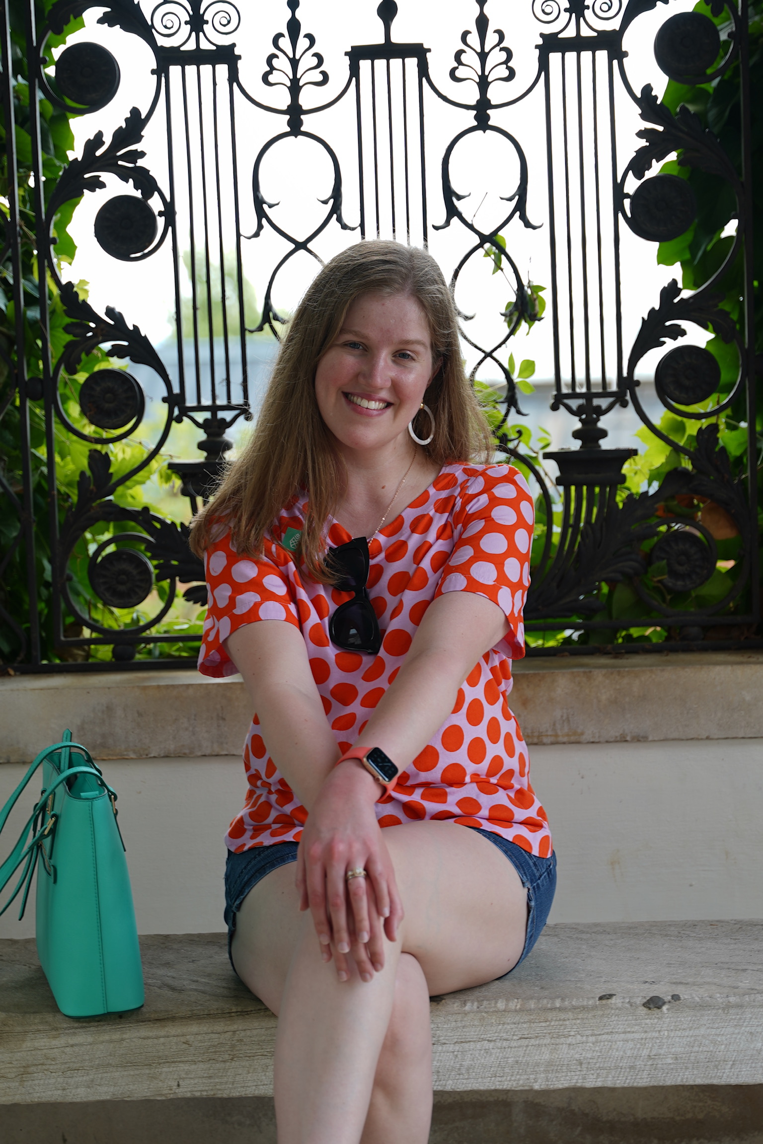 A woman in a red and white polka dot shirt sits on a bench with her legs crossed, smiling, in front of an ornate black iron gate. A turquoise handbag is beside her.