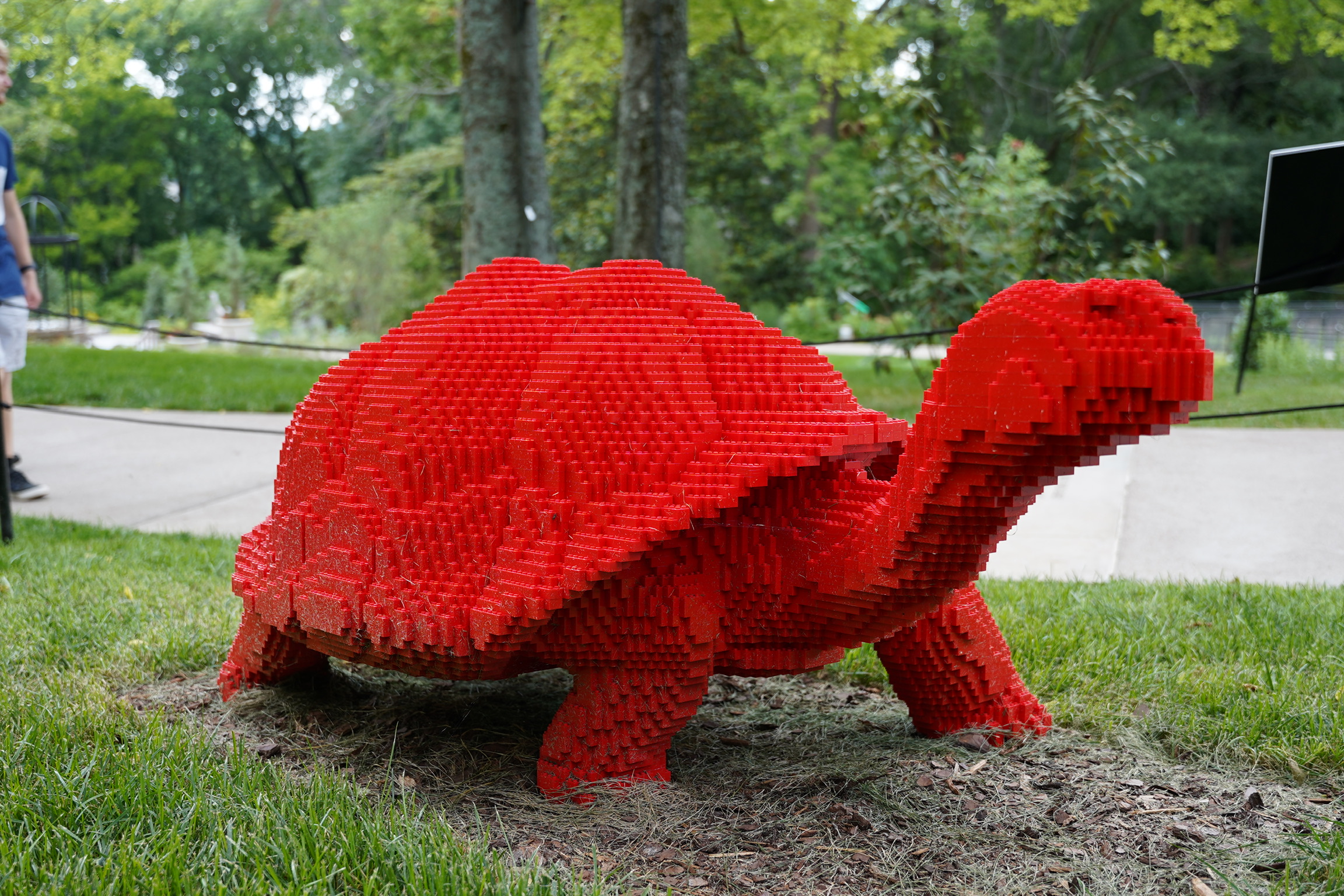 A large red tortoise sculpture made from stacked rectangular blocks is displayed outdoors on grass near a sidewalk and trees.