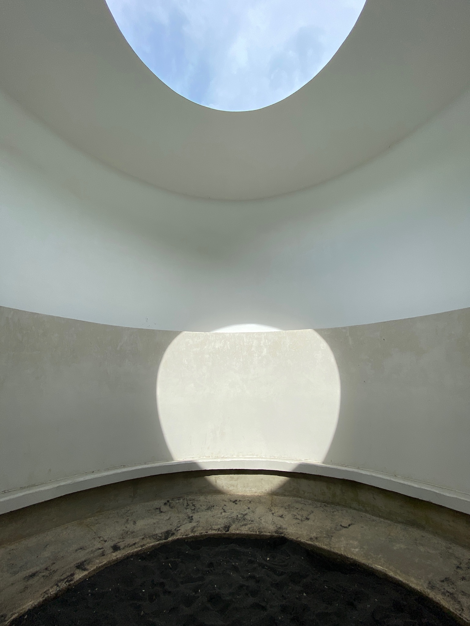 A circular skylight in a white, curved ceiling casts light and shadow onto a rounded wall and a dark floor below.
