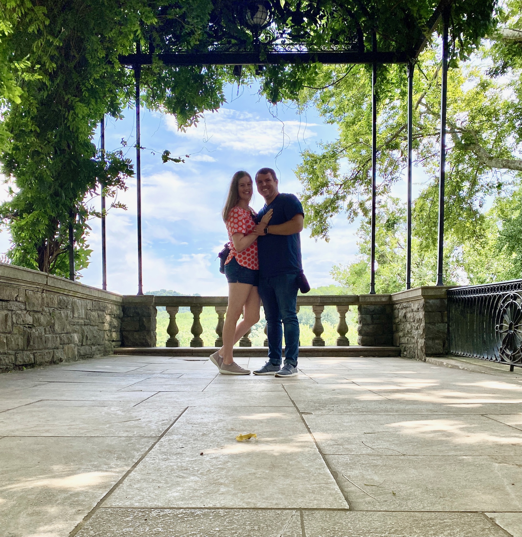 A couple stands close together on a stone patio under a vine-covered pergola, with trees and blue sky in the background.