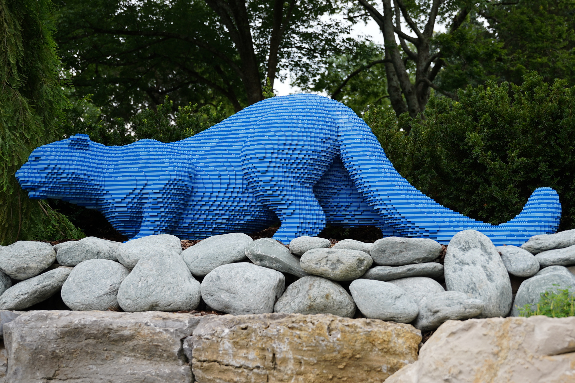 A large blue otter sculpture made of stacked rectangular blocks is displayed outdoors on a stone wall, with trees and bushes in the background.