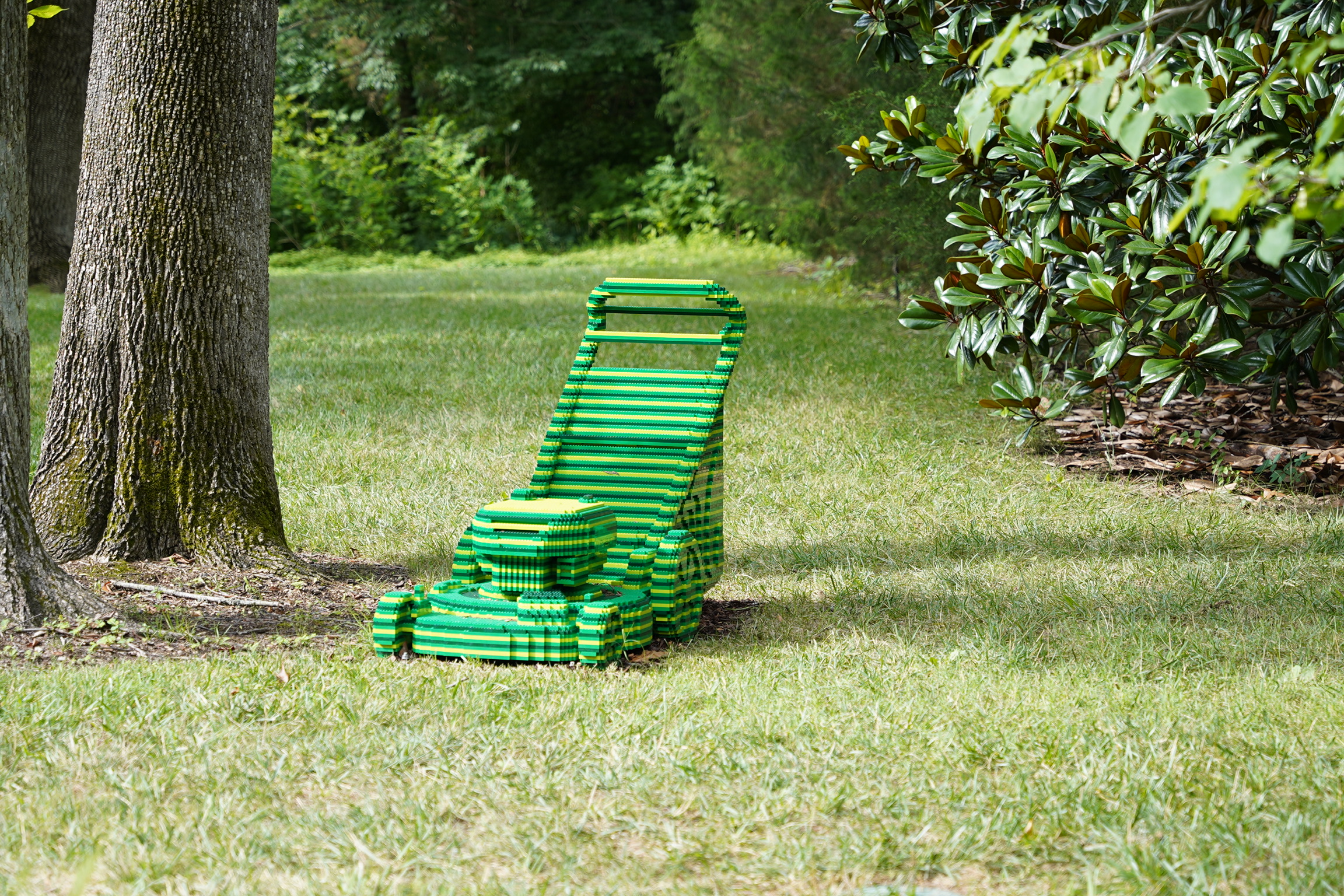 A green and yellow striped lawn mower sits on the grass near several trees in a shaded, leafy backyard.