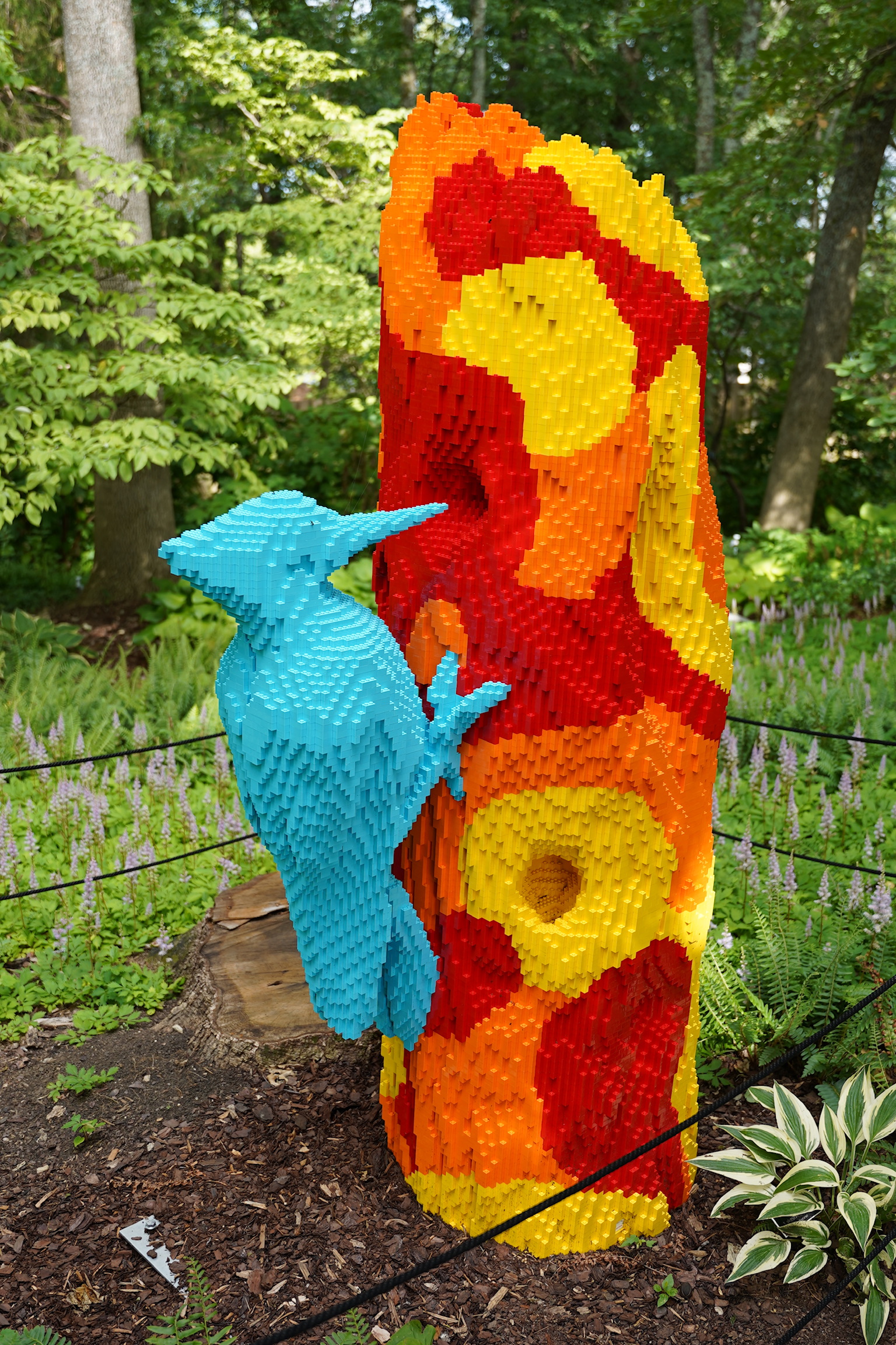 A sculpture of a blue woodpecker on a colorful red, yellow, and orange tree trunk made from LEGO bricks, displayed outdoors in a garden.