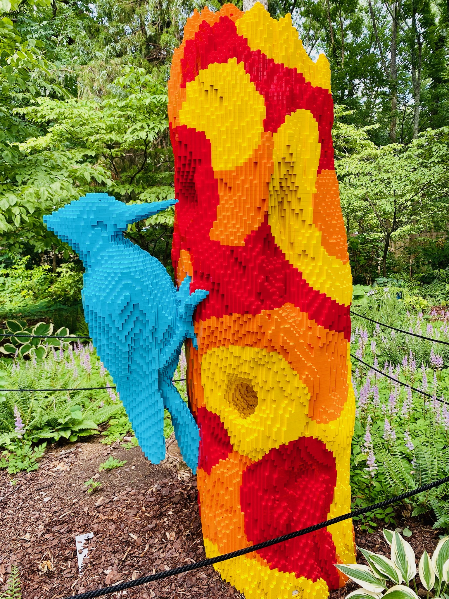 A large sculpture of a blue woodpecker on a colorful red, orange, and yellow tree trunk made from interlocking plastic bricks, set outdoors in a garden.
