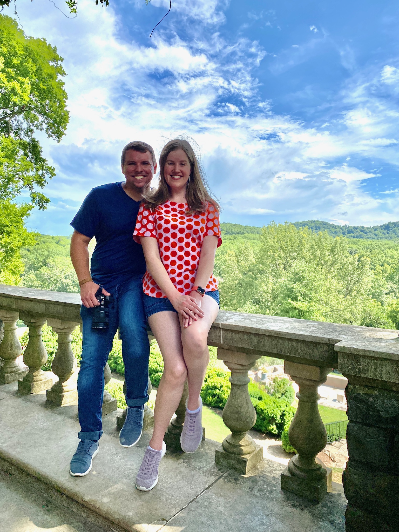A man and woman smile while sitting on a stone railing with green trees and hills in the background under a partly cloudy sky.